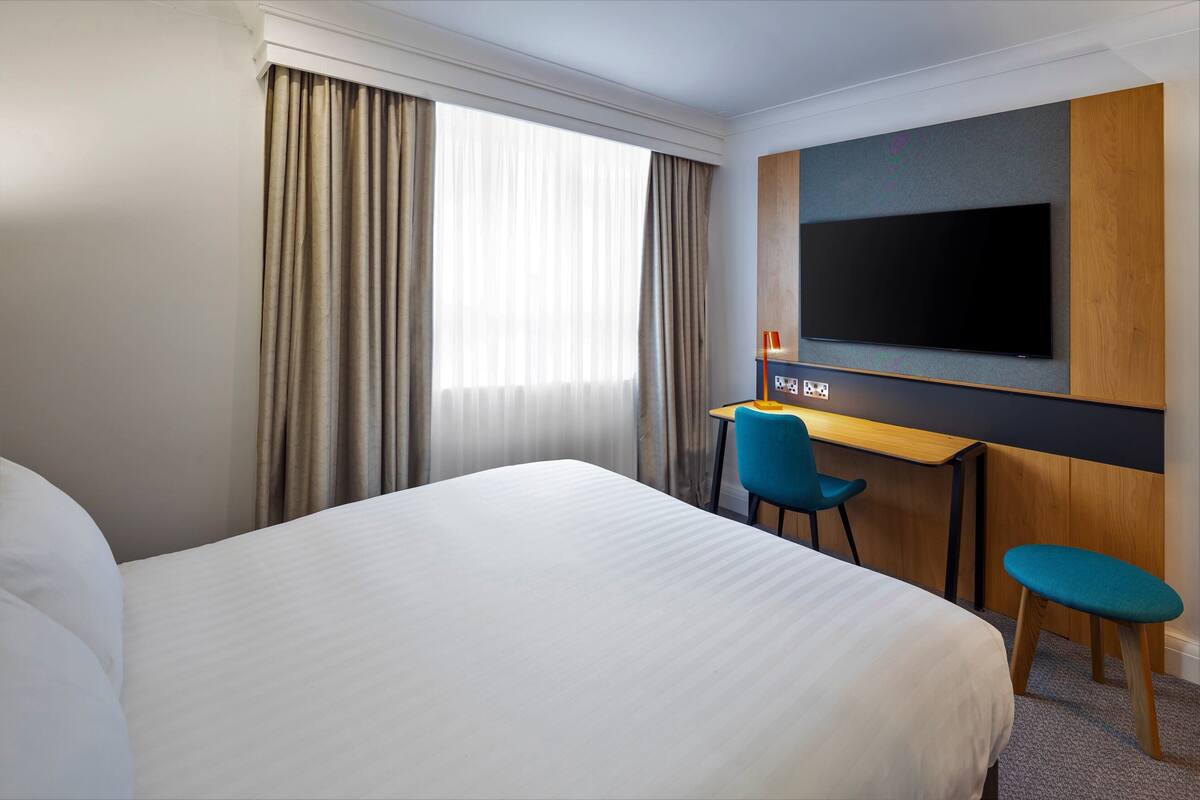 Une chambre d’hôtel avec un grand lit, un bureau avec une chaise, une télévision et une fenêtre avec des rideaux.
