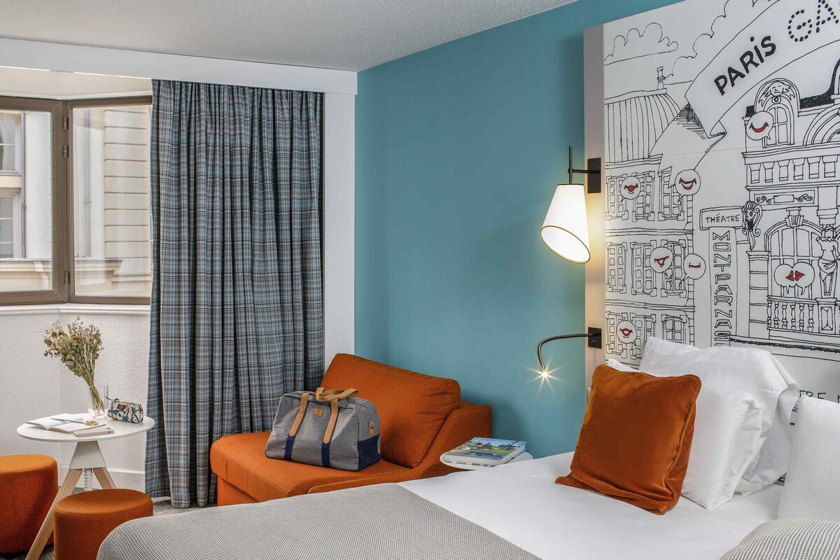 A hotel room with a bed, a seating area with a table, a lamp, and a wall with a Paris-themed illustration.