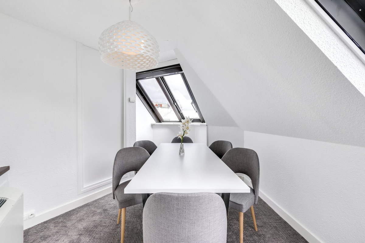 A modern conference room with a white table, grey chairs, and a hanging light fixture.