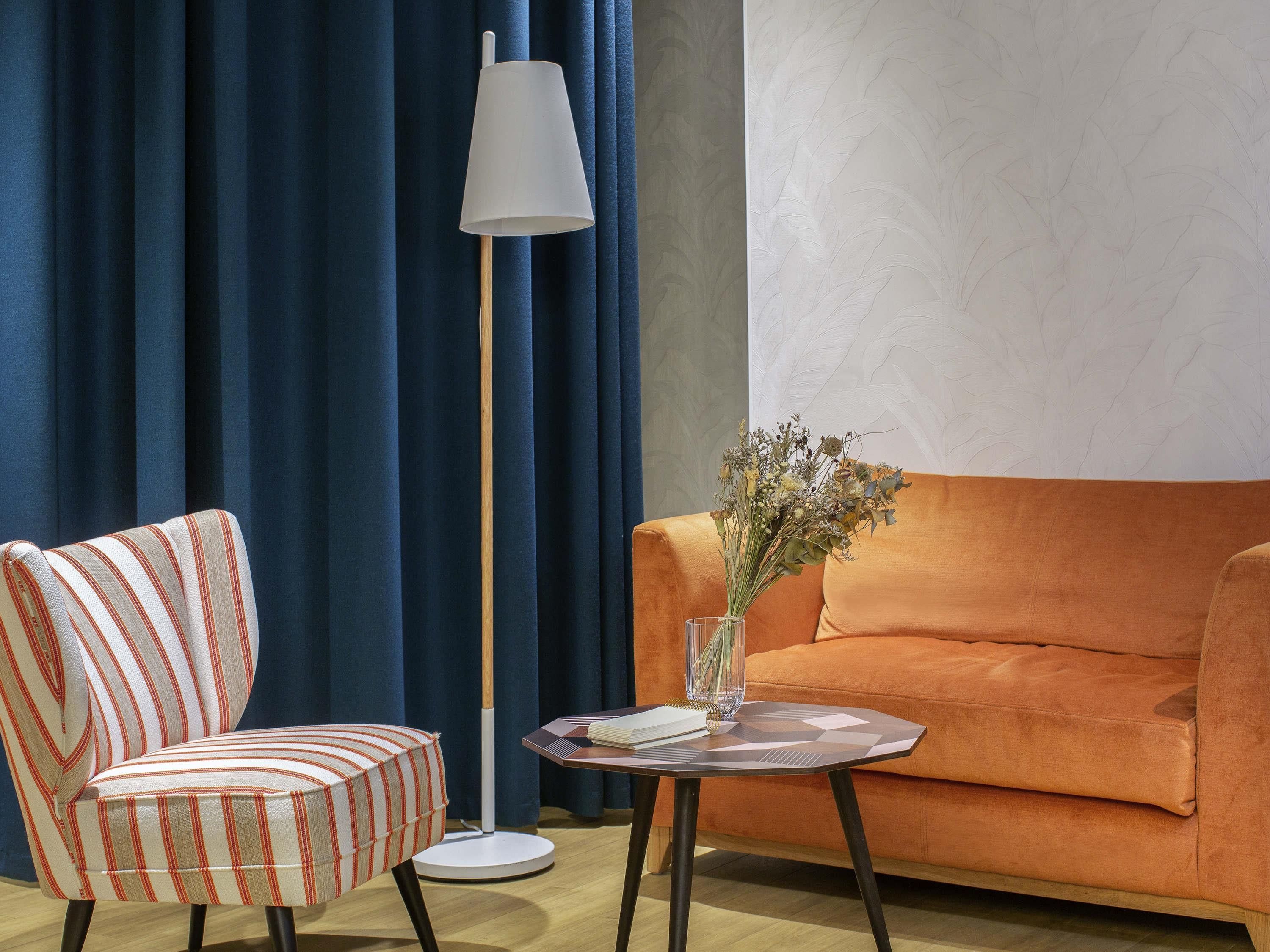 A room with a striped armchair, an orange sofa, a floor lamp, and a small table with a vase of dried flowers.