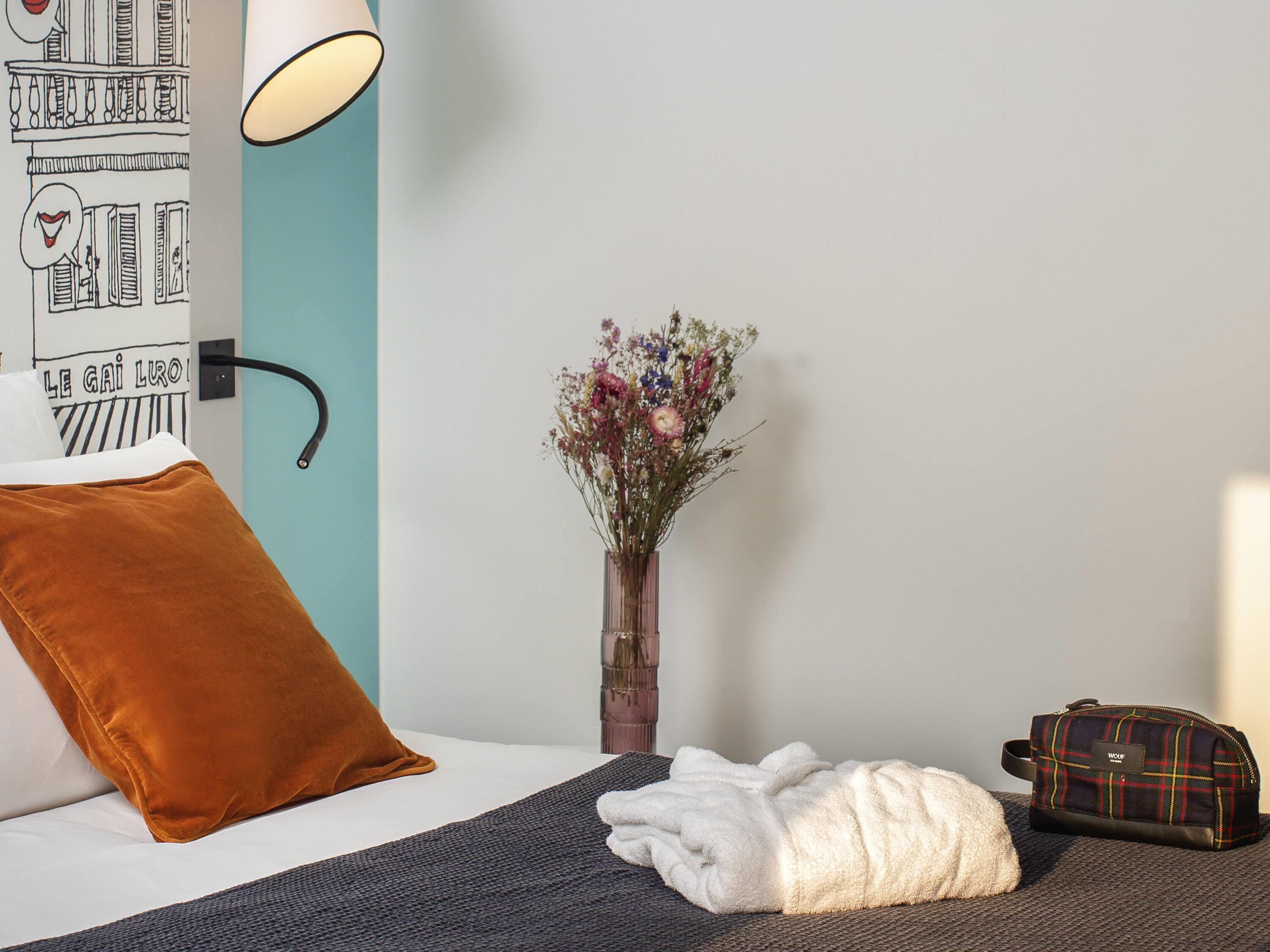 Une chambre d’hôtel avec un lit, une lampe de chevet, un vase avec des fleurs, un oreiller marron, une serviette blanche et un sac à bagages.