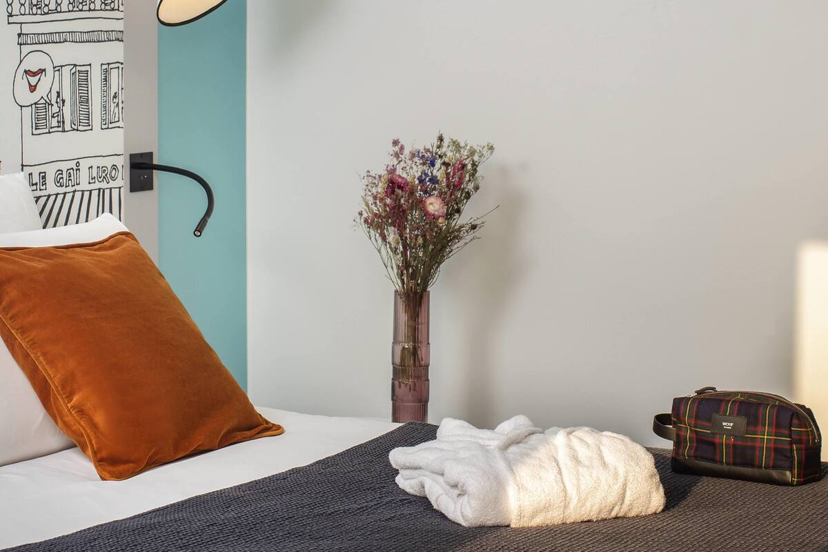 Ett hotellrum med en säng, en sänglampa, en vas med blommor, en brun kudde, en vit handduk och en resväska.