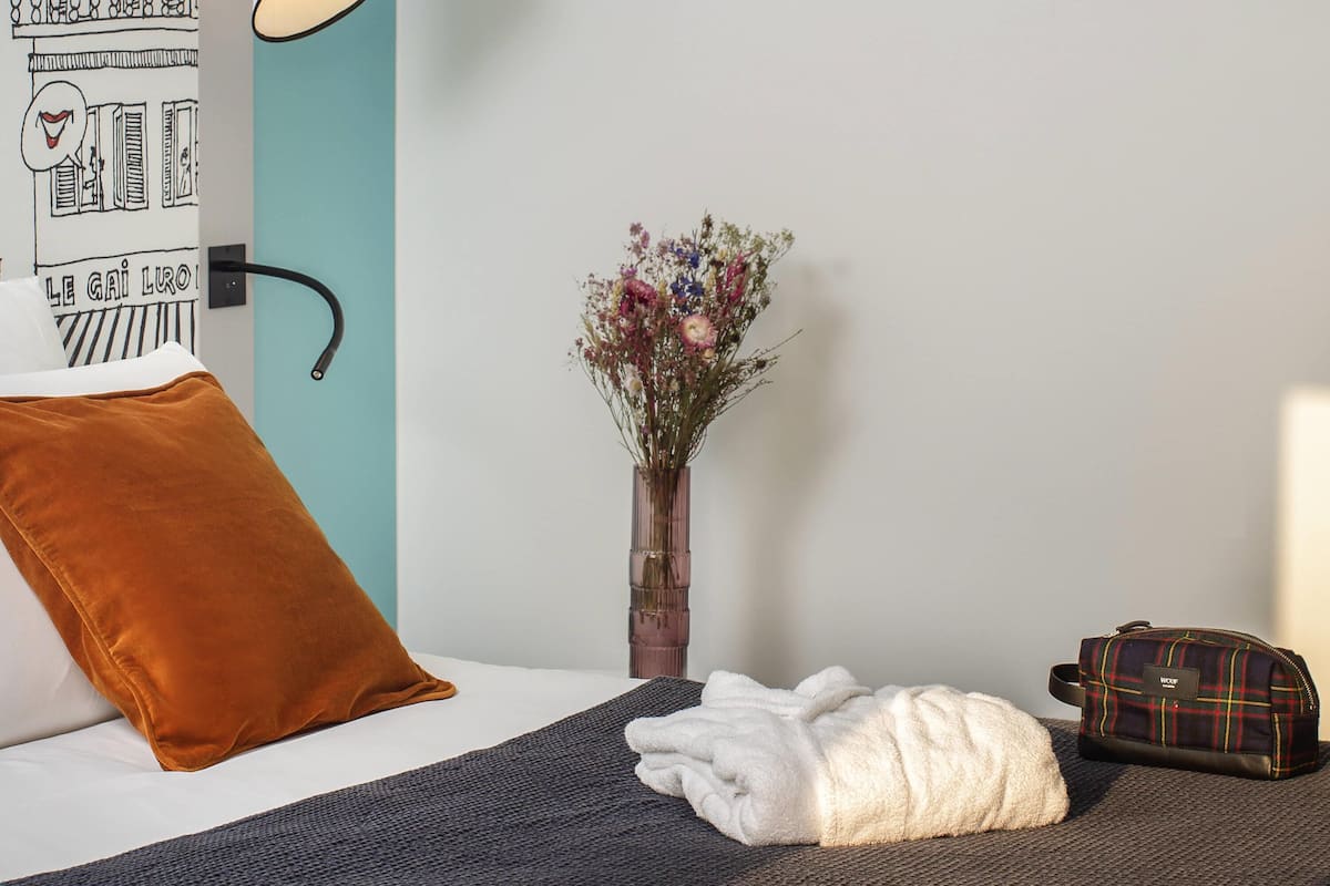 Une chambre d’hôtel avec un lit, une lampe de chevet, un vase avec des fleurs, un oreiller marron, une serviette blanche et un sac à bagages.