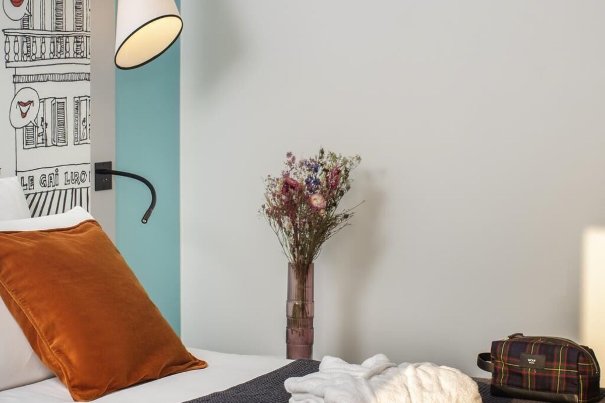 A hotel room with a bed, a brown pillow, a white towel, a small vase with flowers, and a plaid bag.