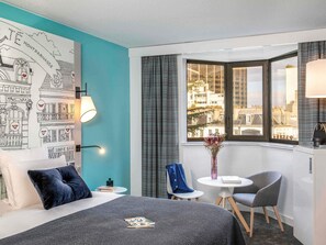 Una habitación de hotel con una cama, un escritorio, una silla, una mesita, un ventanal con vistas a la ciudad y una pared decorada con un mural.