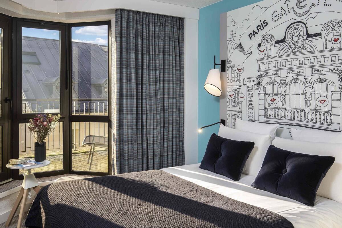 Ein Hotelzimmer mit Bett, Fenster mit Vorhängen, Wand mit einem Wandgemälde zum Thema Paris und einem kleinen Tisch mit Blumenvase.