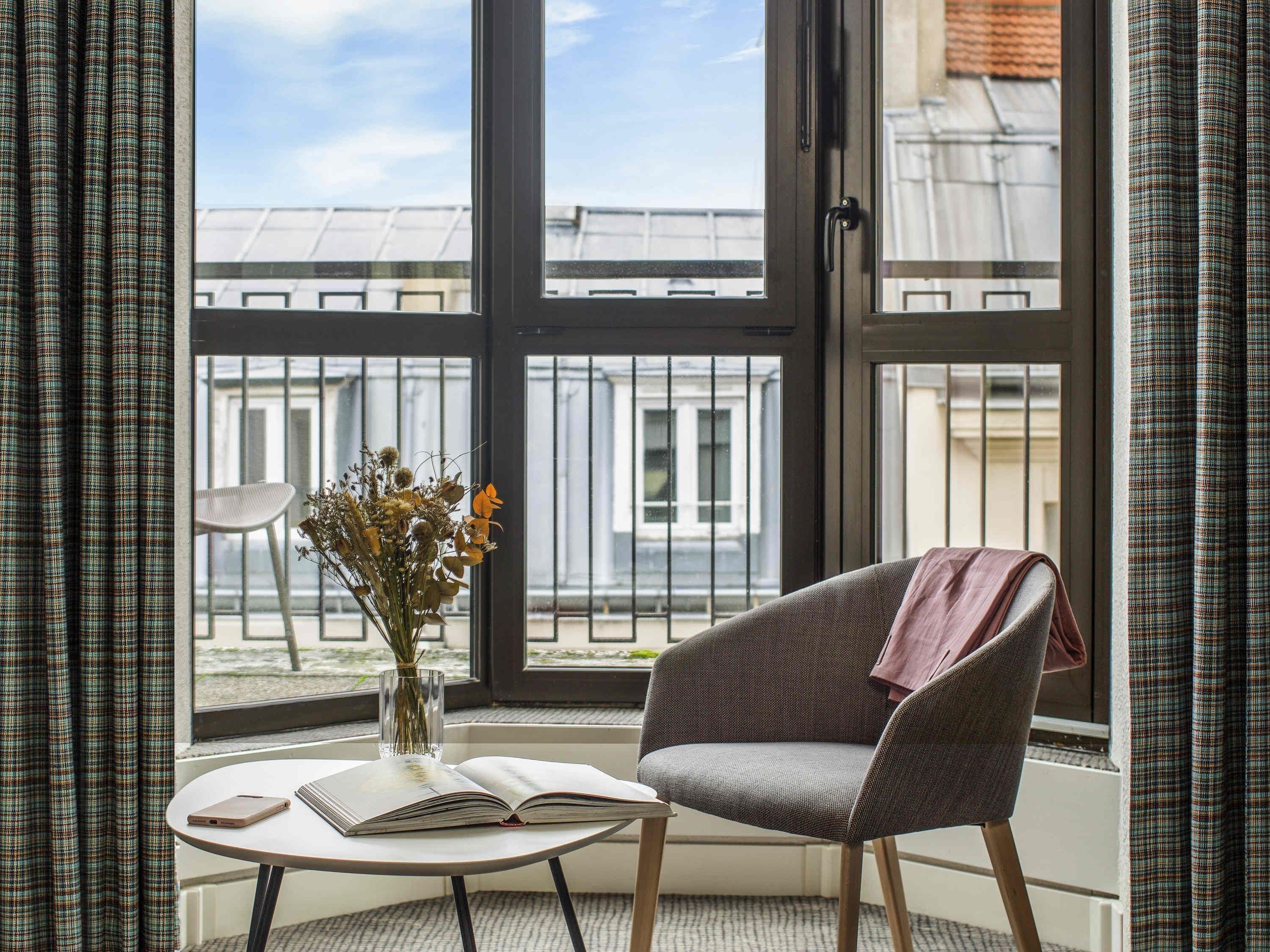 A room with a large window, a chair, a table with a book and a vase of flowers.