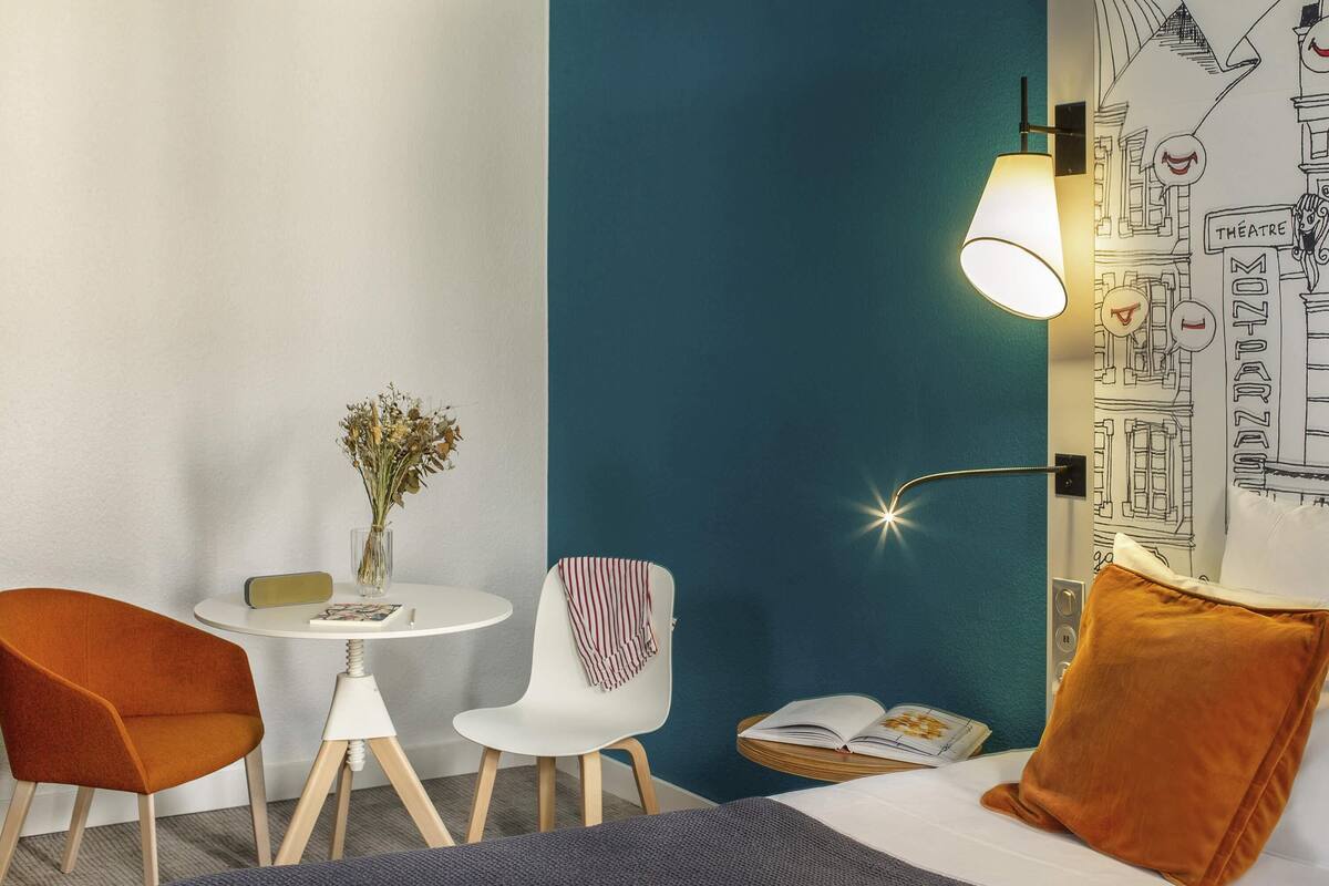 Ein Zimmer mit einem Bett, einem Tisch mit Blumenvase, zwei Stühlen, einer Lampe und einer Wand mit Stadtbild-Zeichnung.
