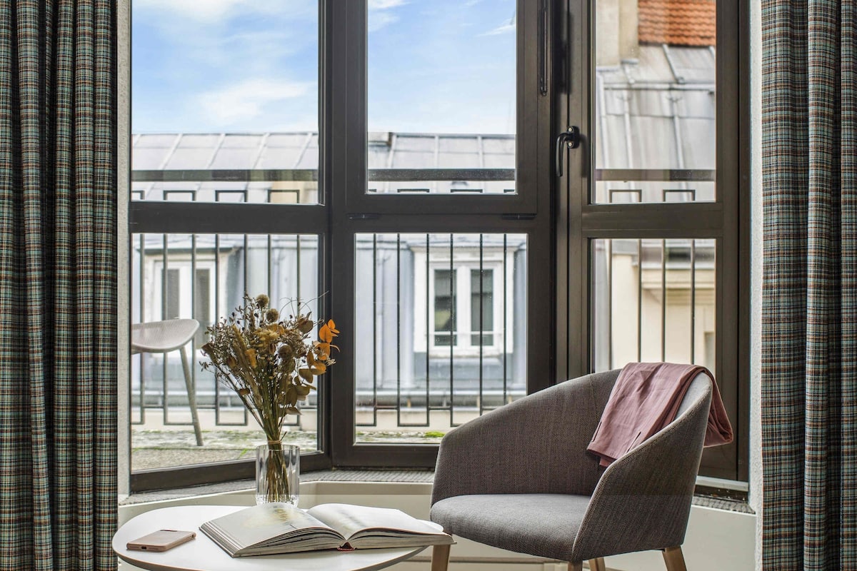 A room with a large window, a chair, a table with a book and a vase of flowers.