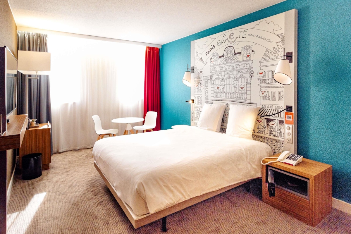 A hotel room with a bed, a desk, a chair, a small table, a lamp, and a wall with a map.