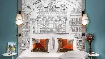 Ein Schlafzimmer mit Wandteppich zum Pariser Thema, zwei Nachttischlampen, einem Bett mit gemusterten Kissen, einem kleinen Tisch mit Buch und Tasse sowie einer Vase mit Blumen.