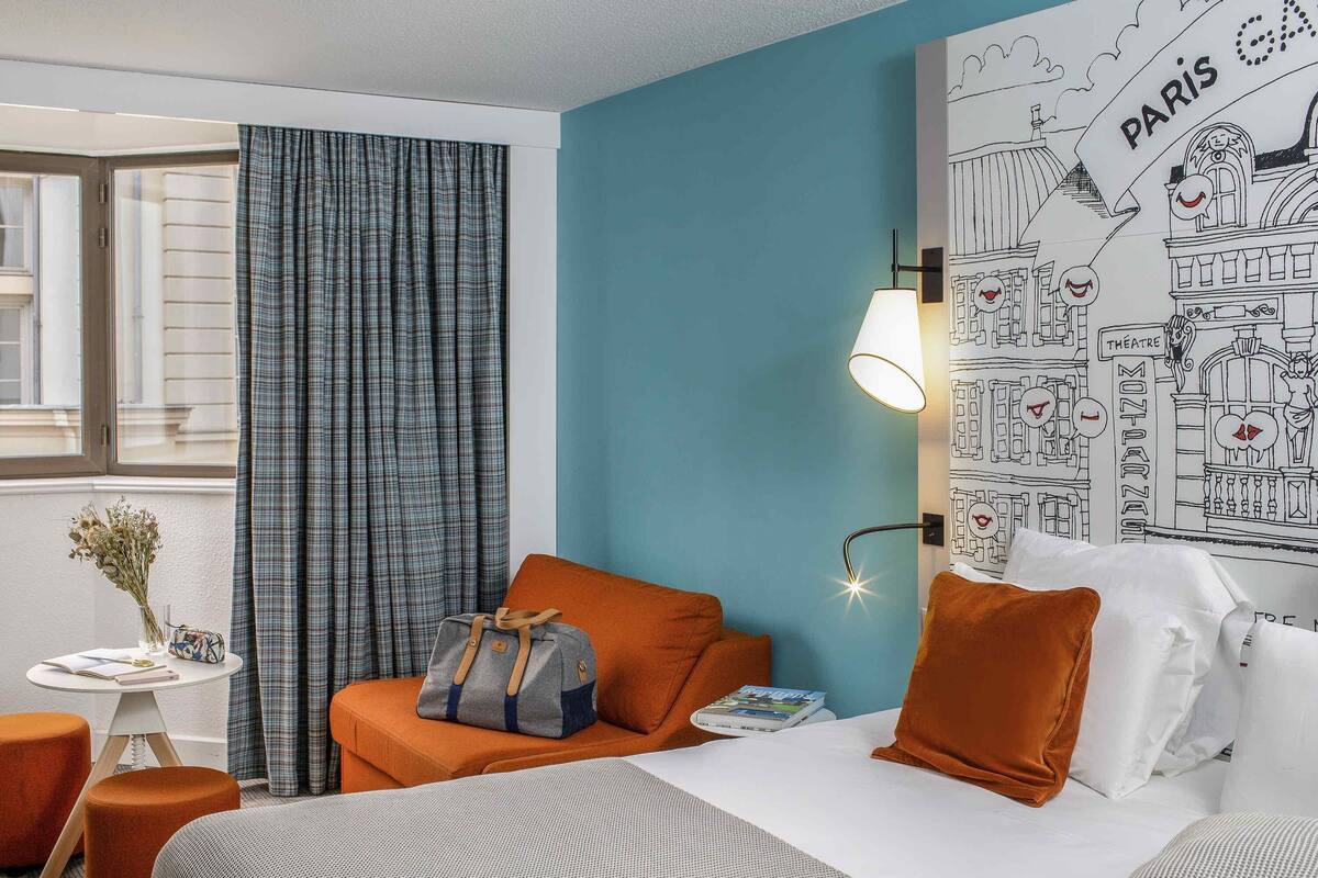Ein Hotelzimmer mit Bett, orangenen Kissen, einer grauen Tasche, einer Lampe, einer Wand mit einer Paris-Karte und einem Fenster mit Vorhängen.