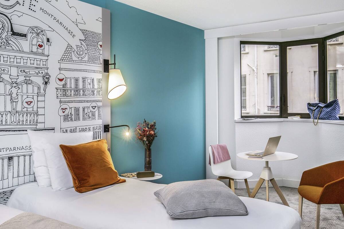 Hotelkamer met een bed, een bureau met een laptop, een stoel, een vaas met bloemen en een muur met een stadsgezicht muurschildering.