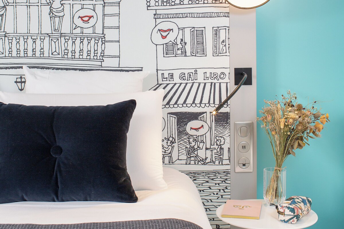 A hotel room with a bed, a wall mural, a lamp, and a small table with a vase and a notebook.
