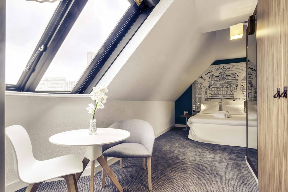 Ein modernes Zimmer mit einem Oberlicht, einem runden Tisch, zwei Stühlen, einem kleinen runden Beistelltisch, einem Bett mit Stadtthema und einem Spiegel.