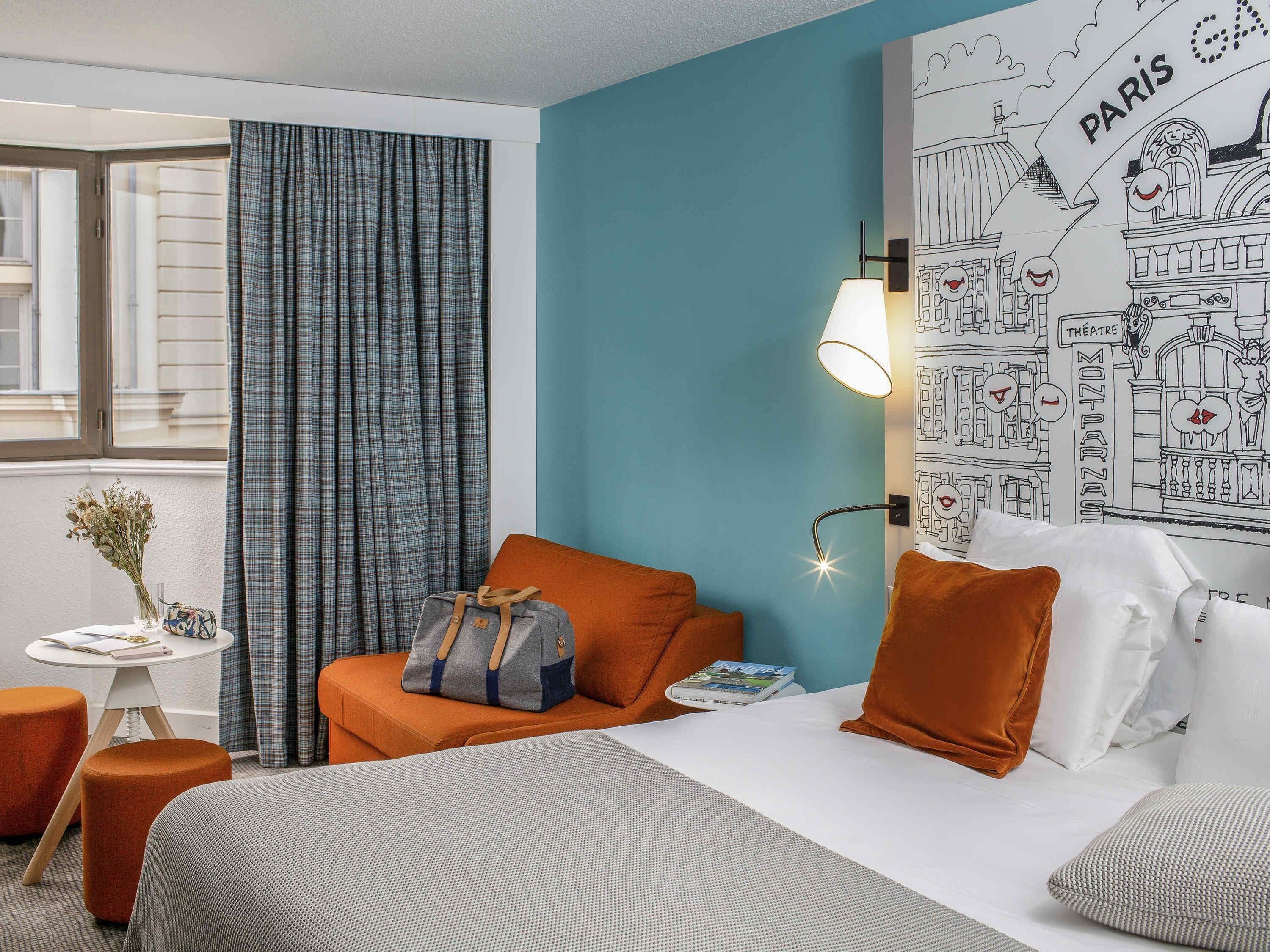 A hotel room with a bed, a seating area with a table, a lamp, and a wall with a Paris-themed illustration.