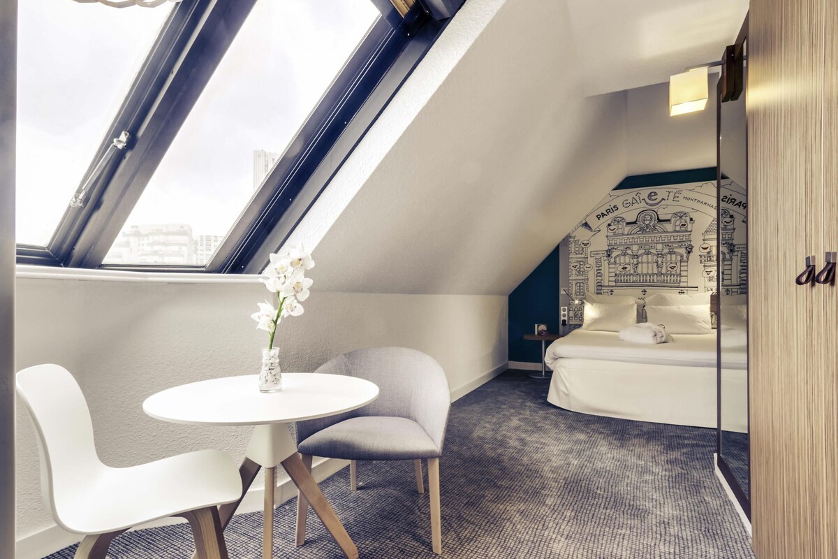 A modern room with a skylight, a round table, two chairs, a small round table, a bed with a city-themed headboard, and a mirror.