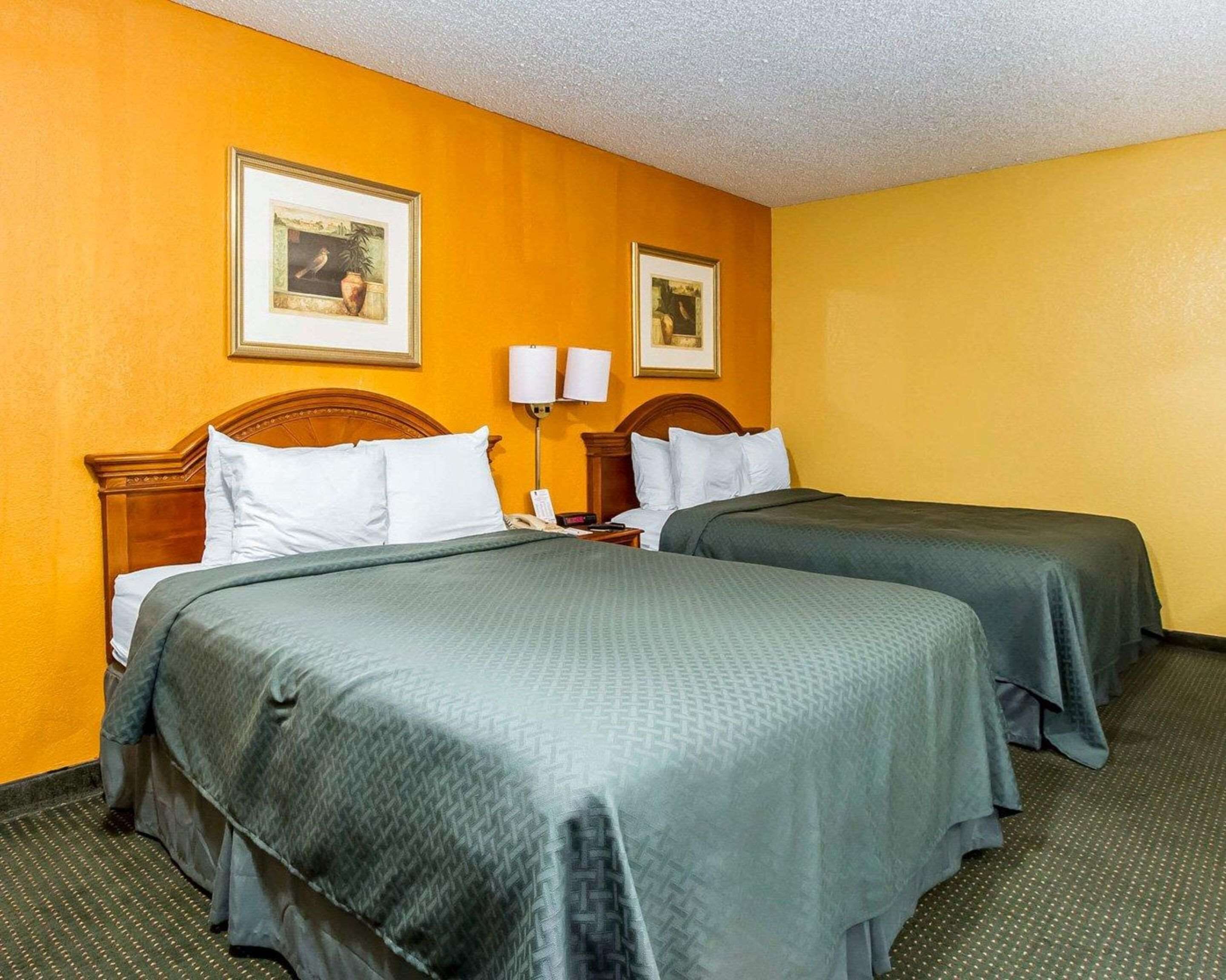 A hotel room with two beds, each with a green bedspread, white pillows, and a wooden headboard. There are two framed pictures on the wall above the beds.