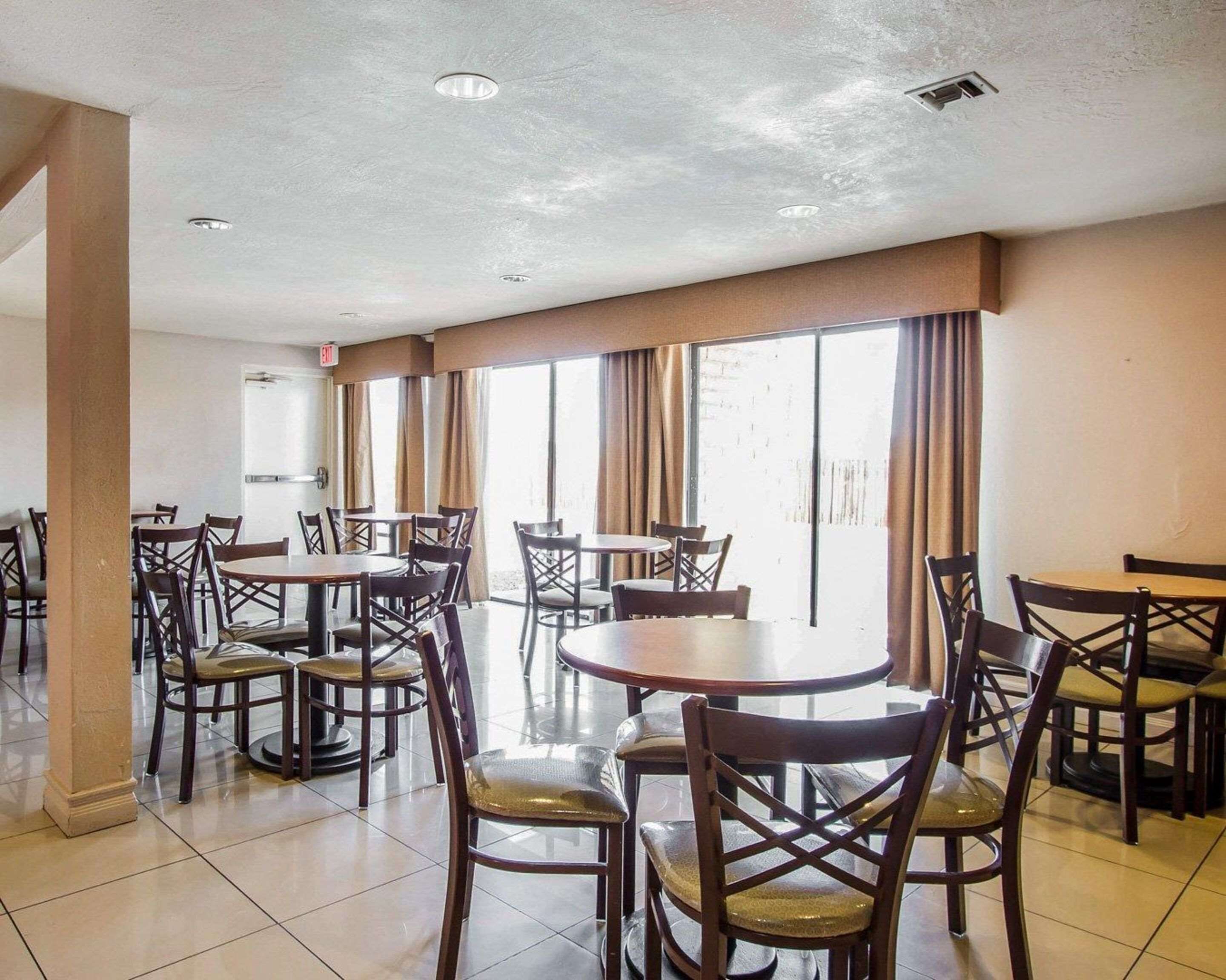 A dining area with multiple round tables and chairs, large windows with curtains, and a tiled floor.