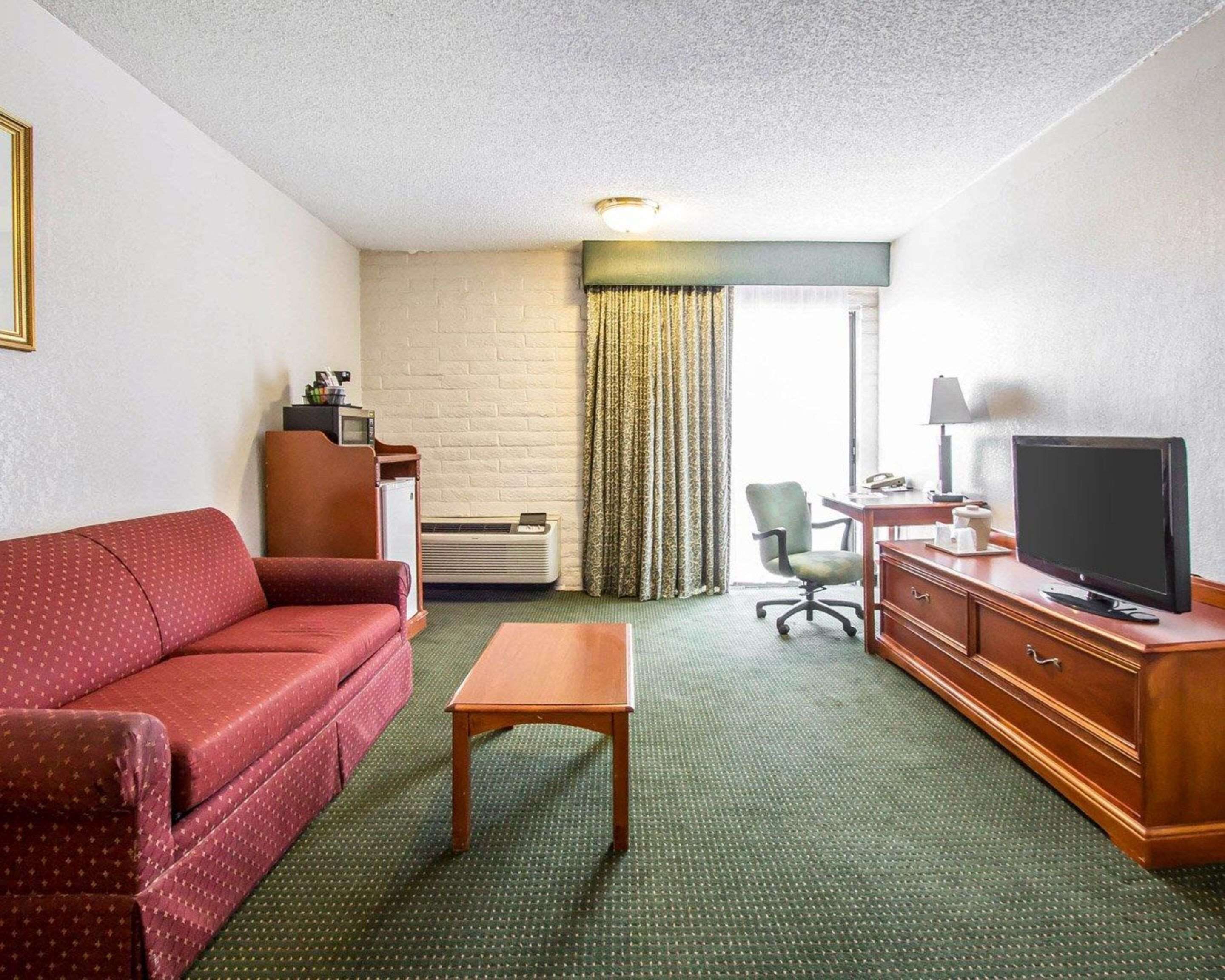 A hotel room with a red sofa, a wooden table, a TV on a wooden cabinet, a desk with a lamp, a chair, and a window with curtains.