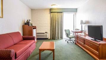 A hotel room with a red sofa, a wooden table, a TV on a wooden cabinet, a desk with a lamp, a chair, and a window with curtains.