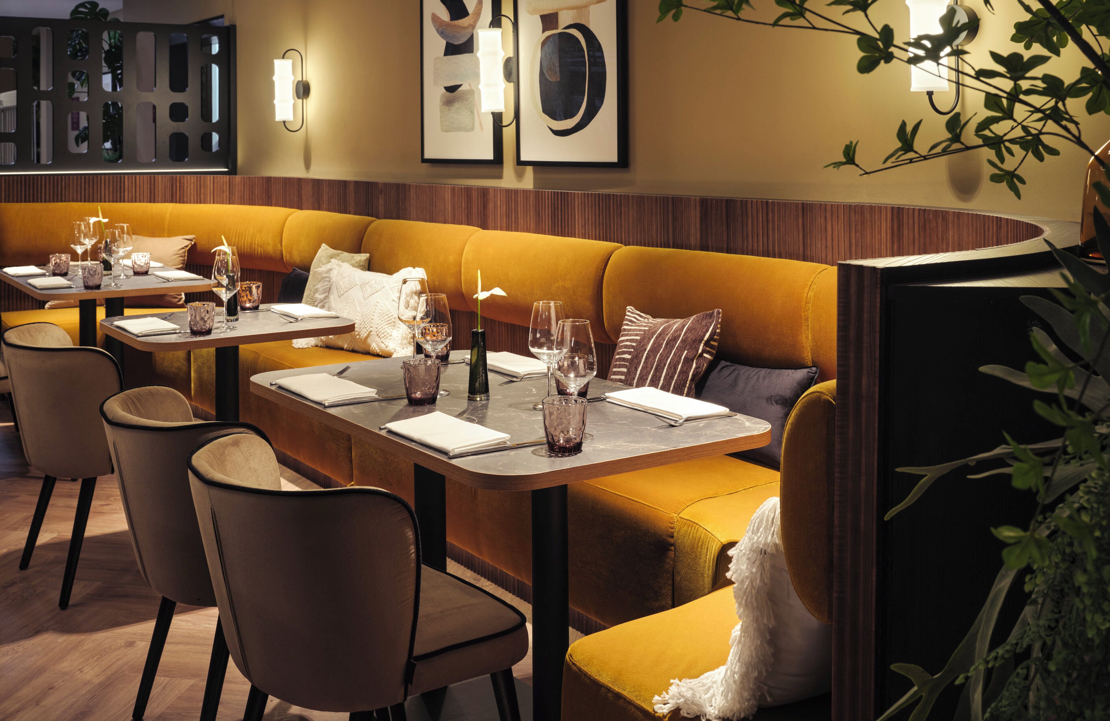 A restaurant with a long, curved wooden bench seating area, multiple tables with chairs, and a warm, inviting ambiance with wall-mounted lighting and framed artwork.