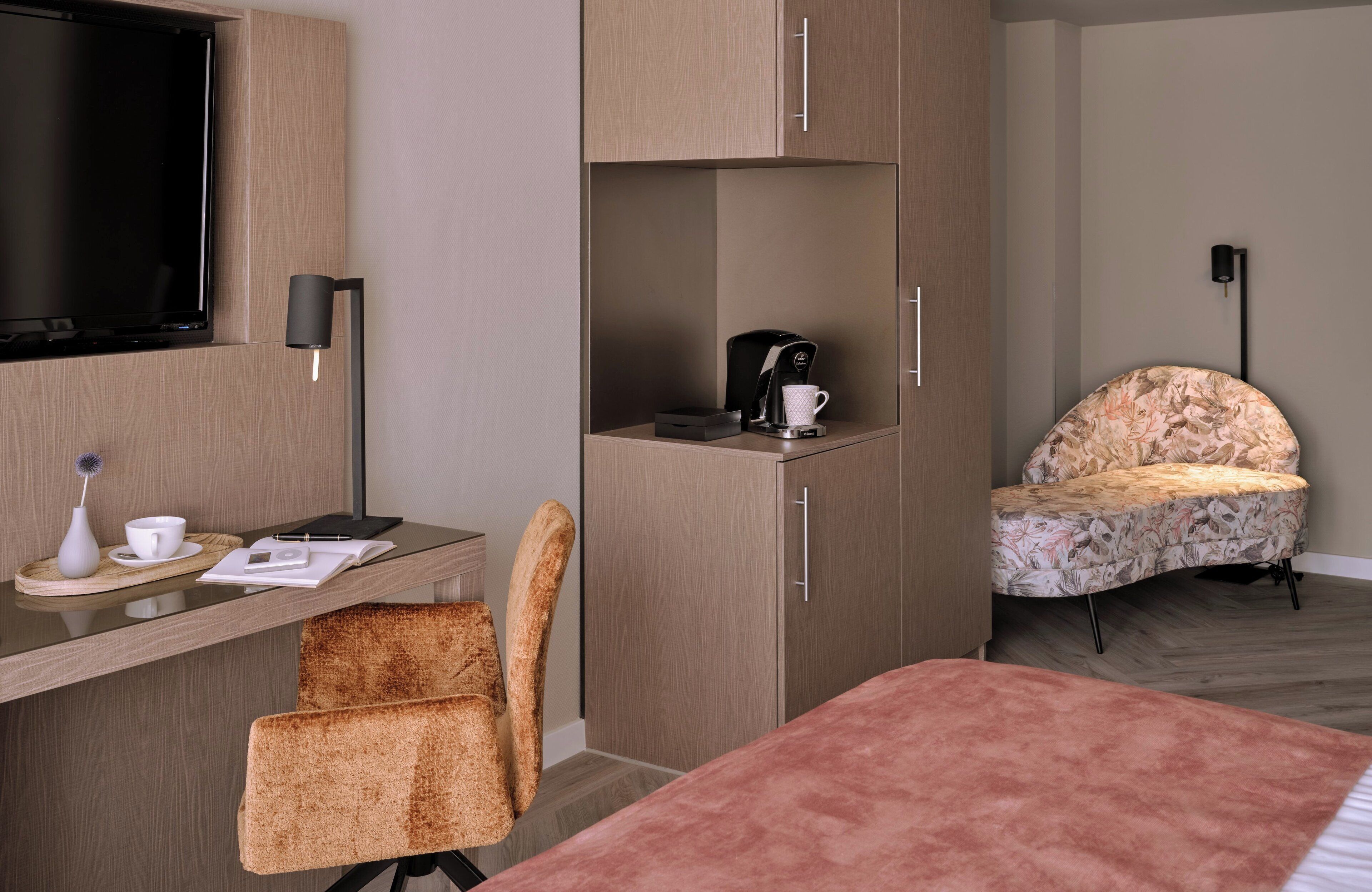 A hotel room with a bed, a desk with a chair, a television, a coffee maker, and a decorative chair.