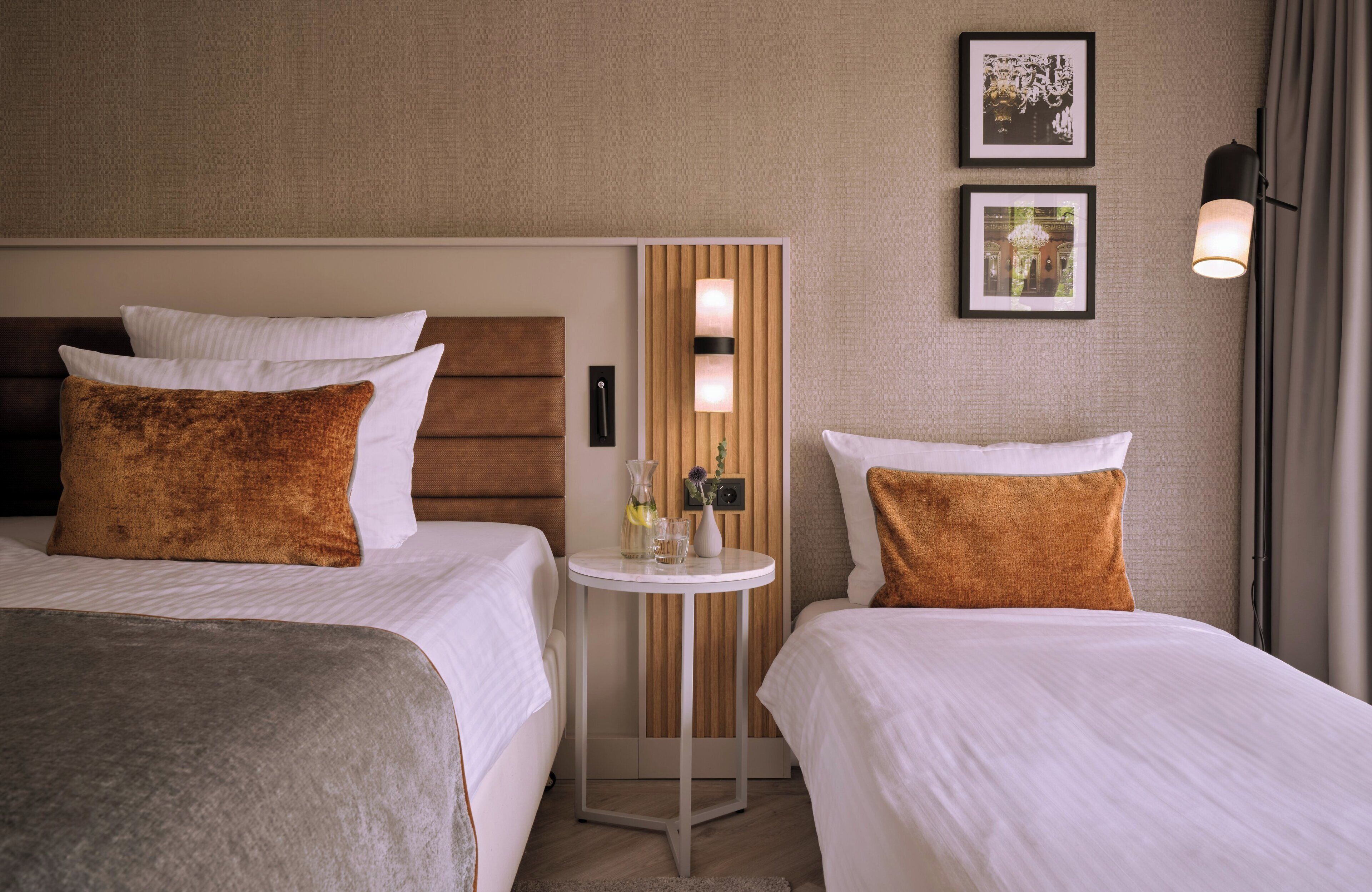 Ein Hotelzimmer mit zwei Betten, einem kleinen runden Tisch mit einer Vase und einer Flasche sowie zwei gerahmten Bildern an der Wand.