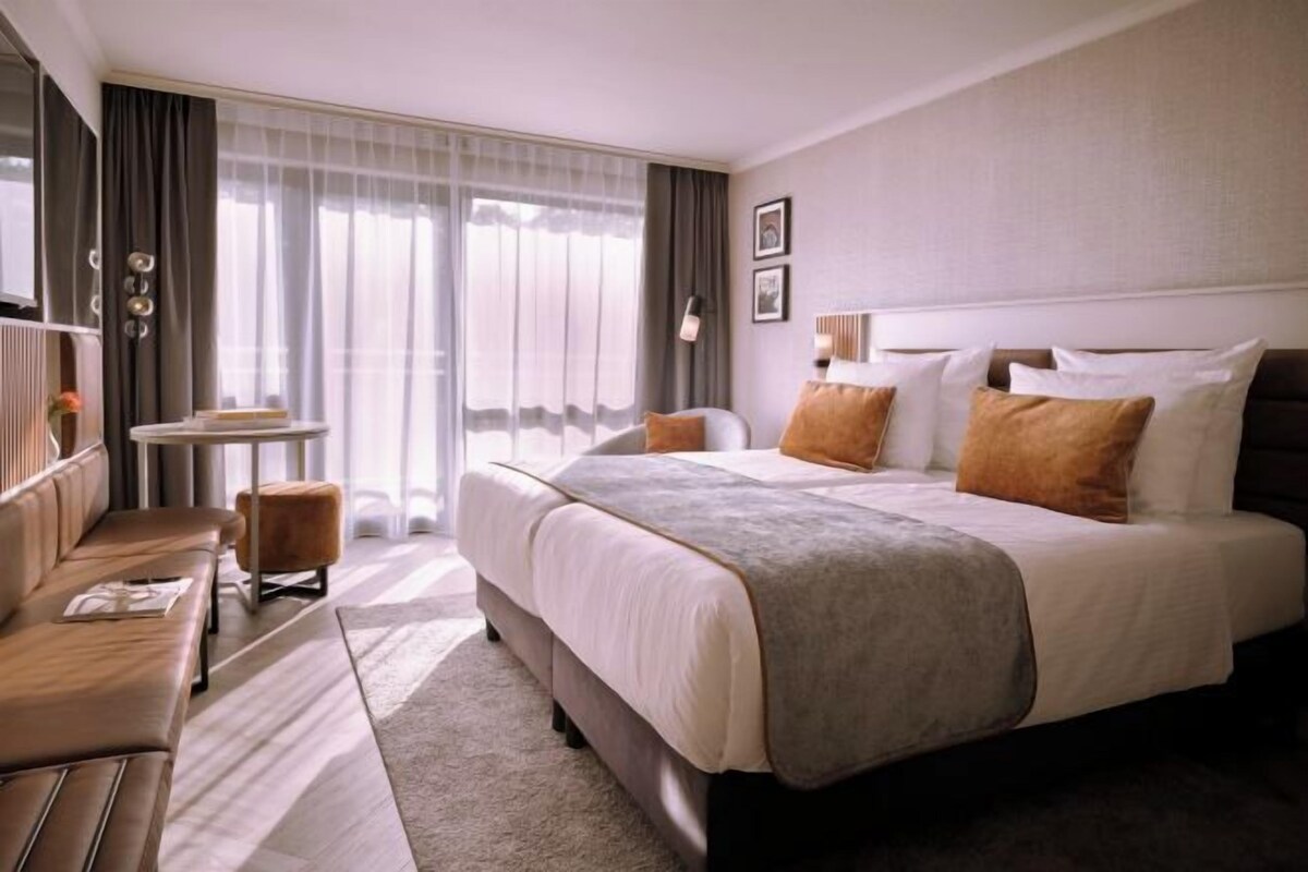 Een hotelkamer met een groot bed, een zithoek met een tafeltje en een groot raam met gordijnen.