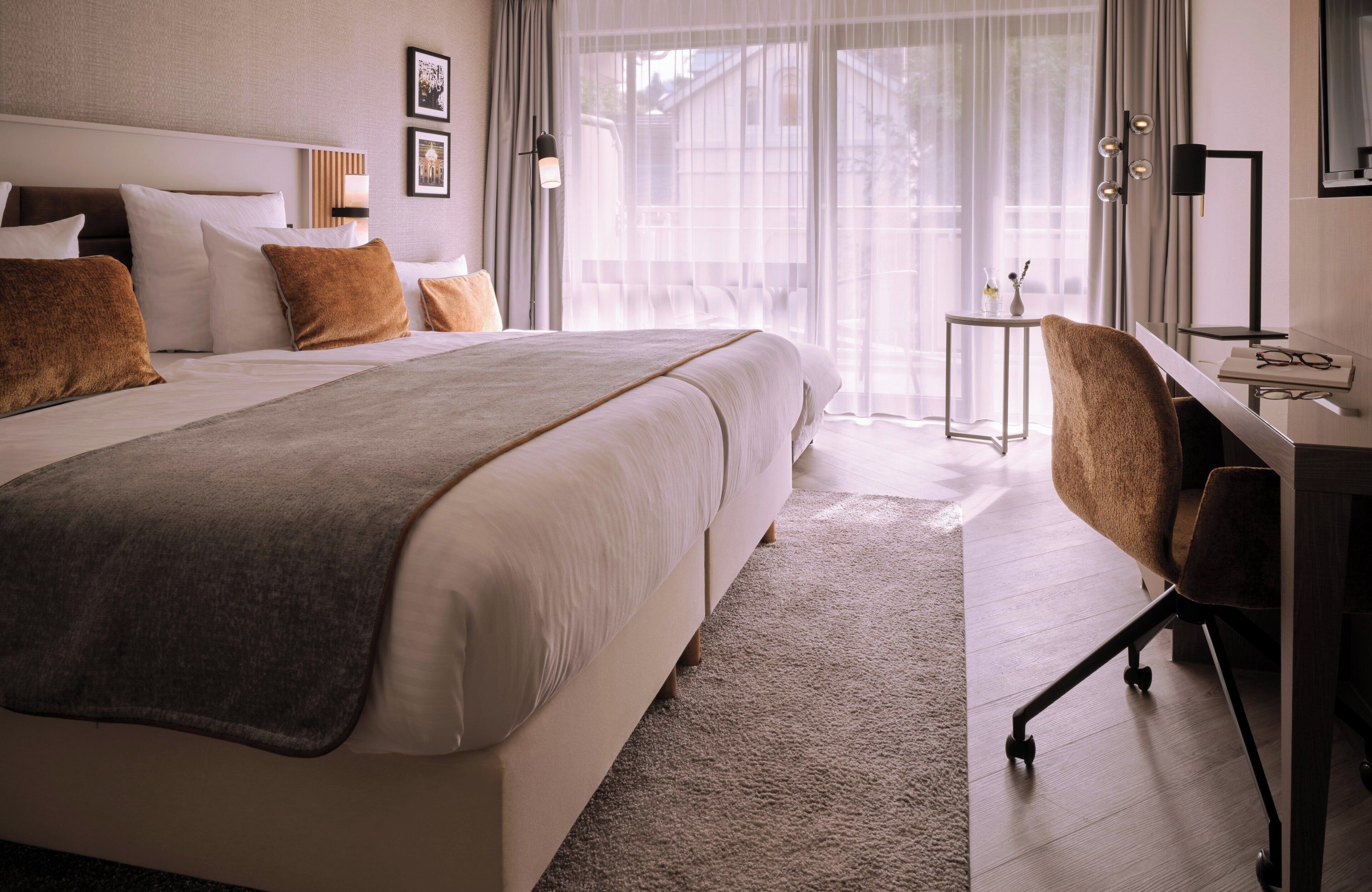 A modern hotel room with a large bed, a desk, and a view of the outdoors through sheer curtains.