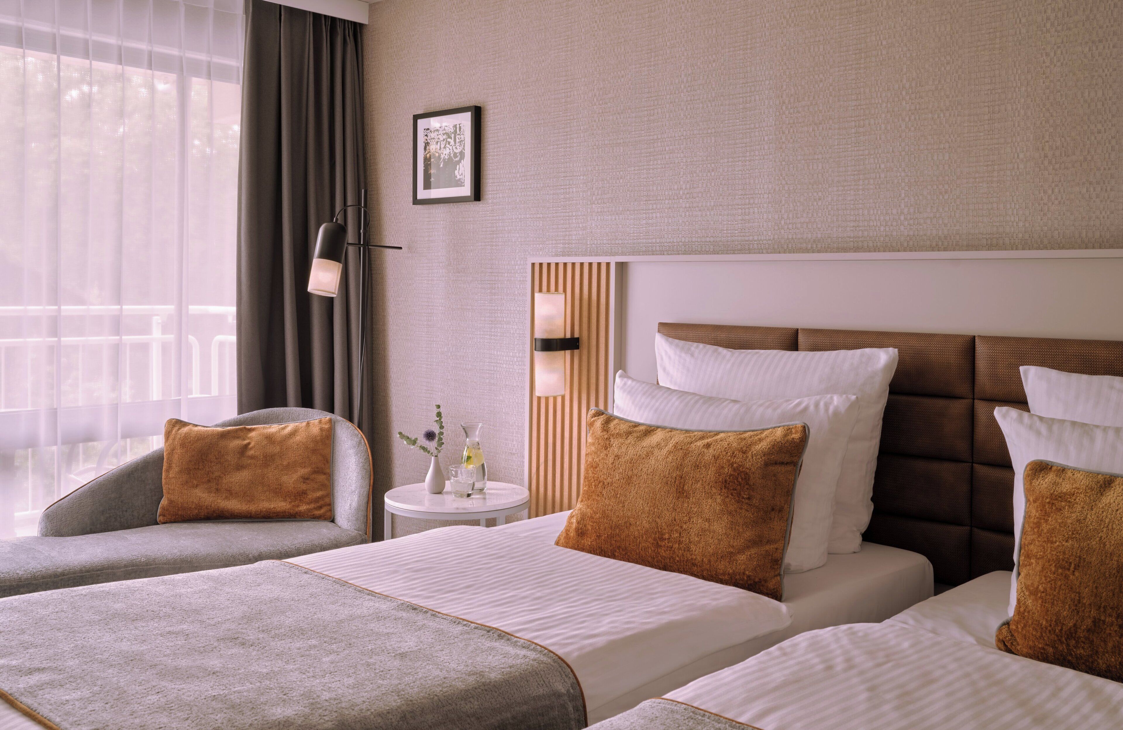 Une chambre d’hôtel avec deux lits, un canapé, une petite table avec un vase et une vue sur l’extérieur à travers des rideaux légers.