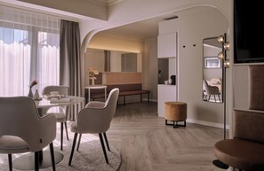 A modern hotel room with a round dining table, two chairs, a bench, a small ottoman, and a large mirror with lights.