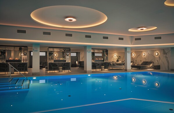 A modern indoor swimming pool with a blue color, surrounded by lounge areas with chairs and tables.