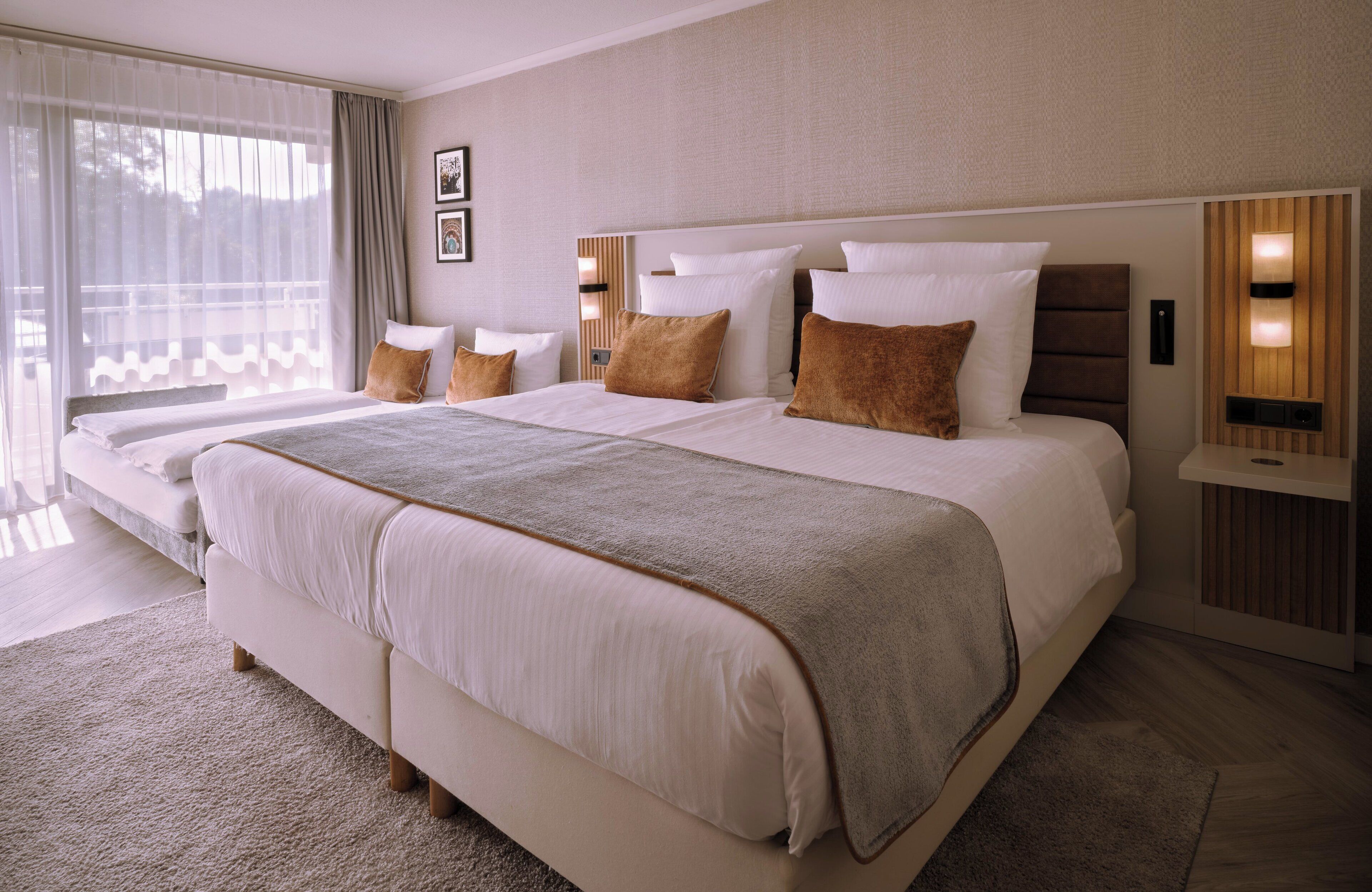 A large bed with multiple pillows, a bedside table with a lamp, and a view of the outdoors through large windows.