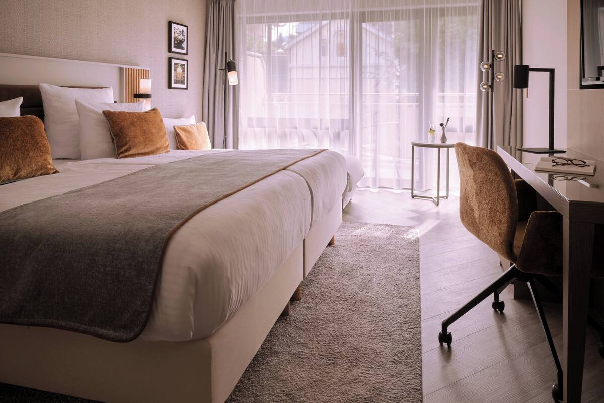 Une chambre d’hôtel moderne, dotée d’un grand lit, d’un bureau et offrant une vue sur l’extérieur à travers de fines rideaux.