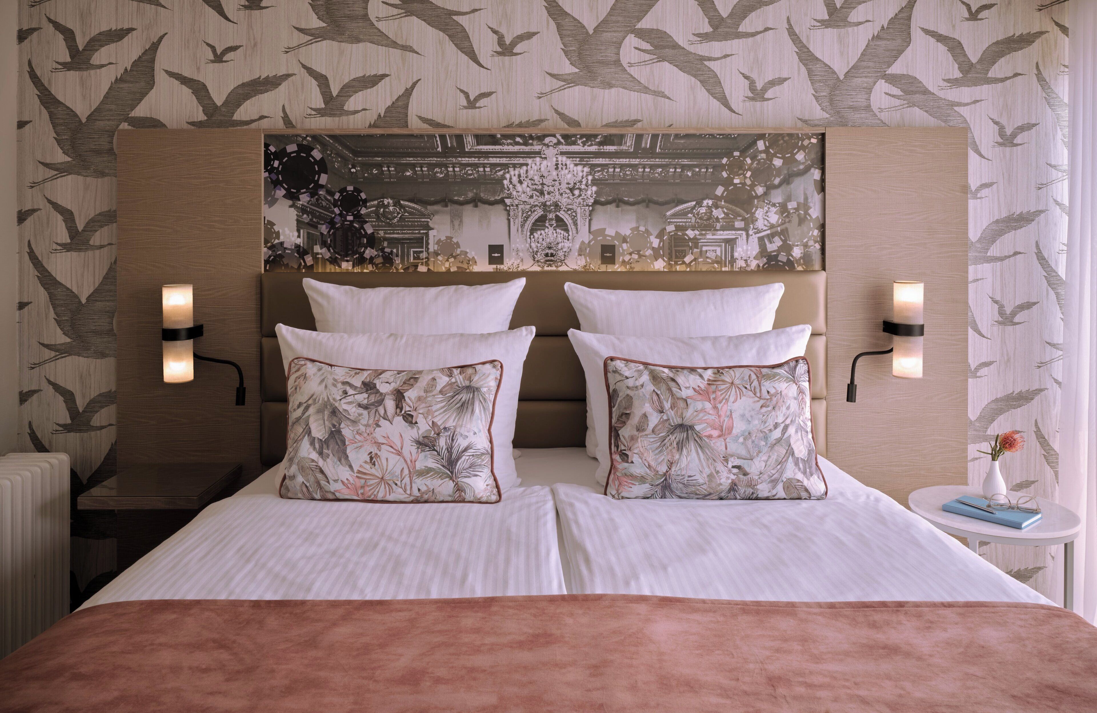 A double bed with floral pillows, a headboard with a decorative mural, and two wall-mounted reading lamps.