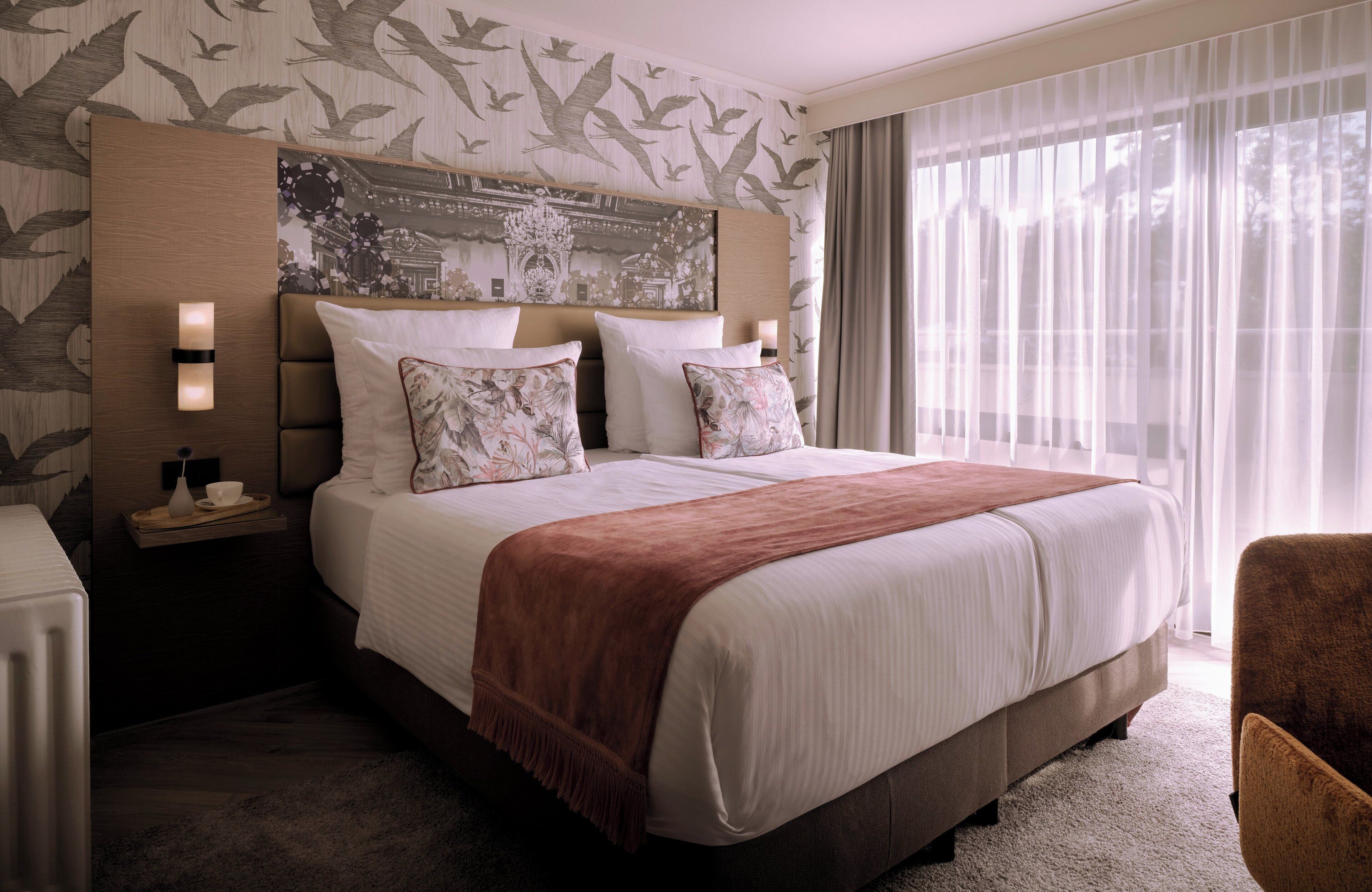 Quarto de hotel com uma cama grande, mesa de cabeceira com abajur e vista para o exterior através de cortinas translúcidas.