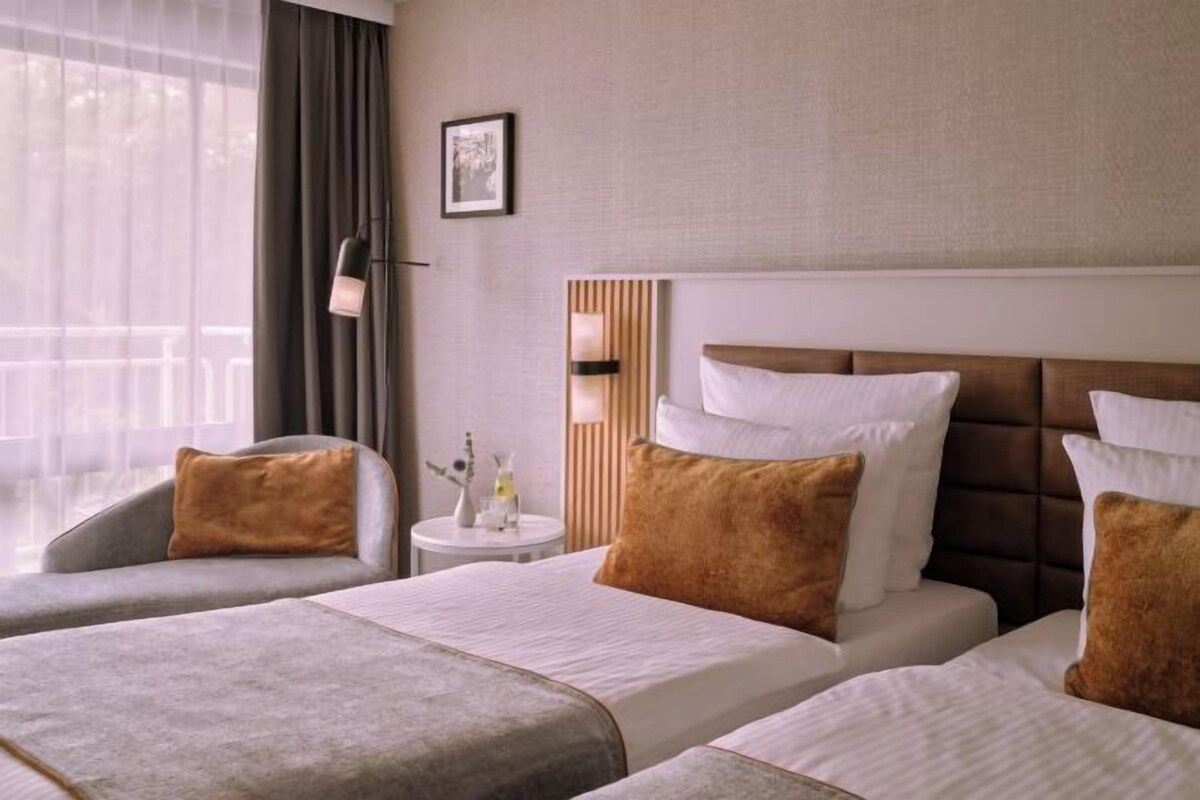 A hotel room with two beds, a sofa, a small round table with a lamp, and a window with curtains.