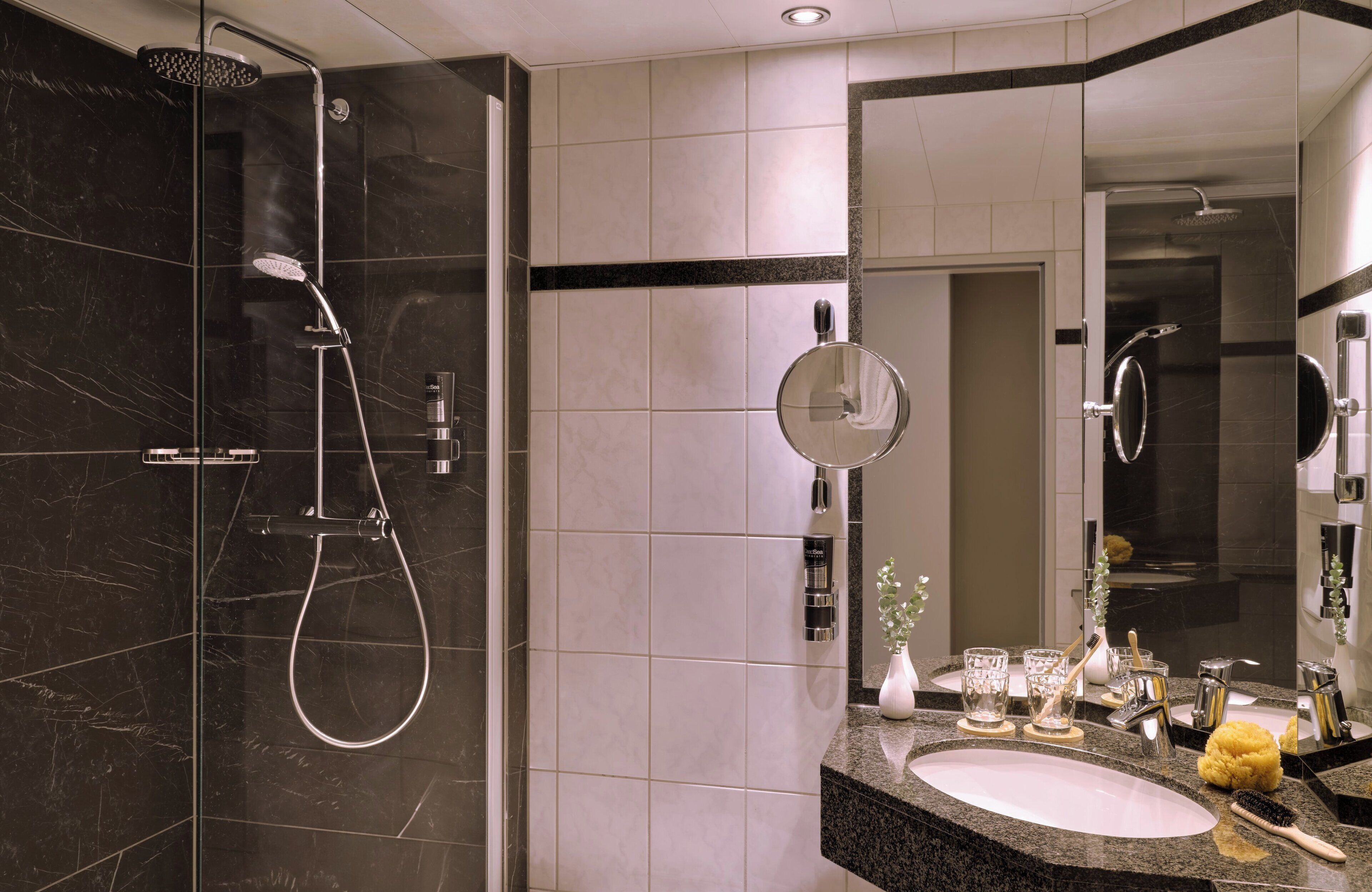 Un baño moderno con ducha envuelta en vidrio, un espejo grande y encimera de granito con lavabo y artículos de aseo.