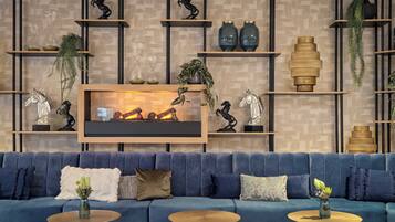 Una sala de estar moderna con un sofá azul, dos mesas redondas de centro y una chimenea. Estantes con objetos decorativos y plantas están montados en la pared.