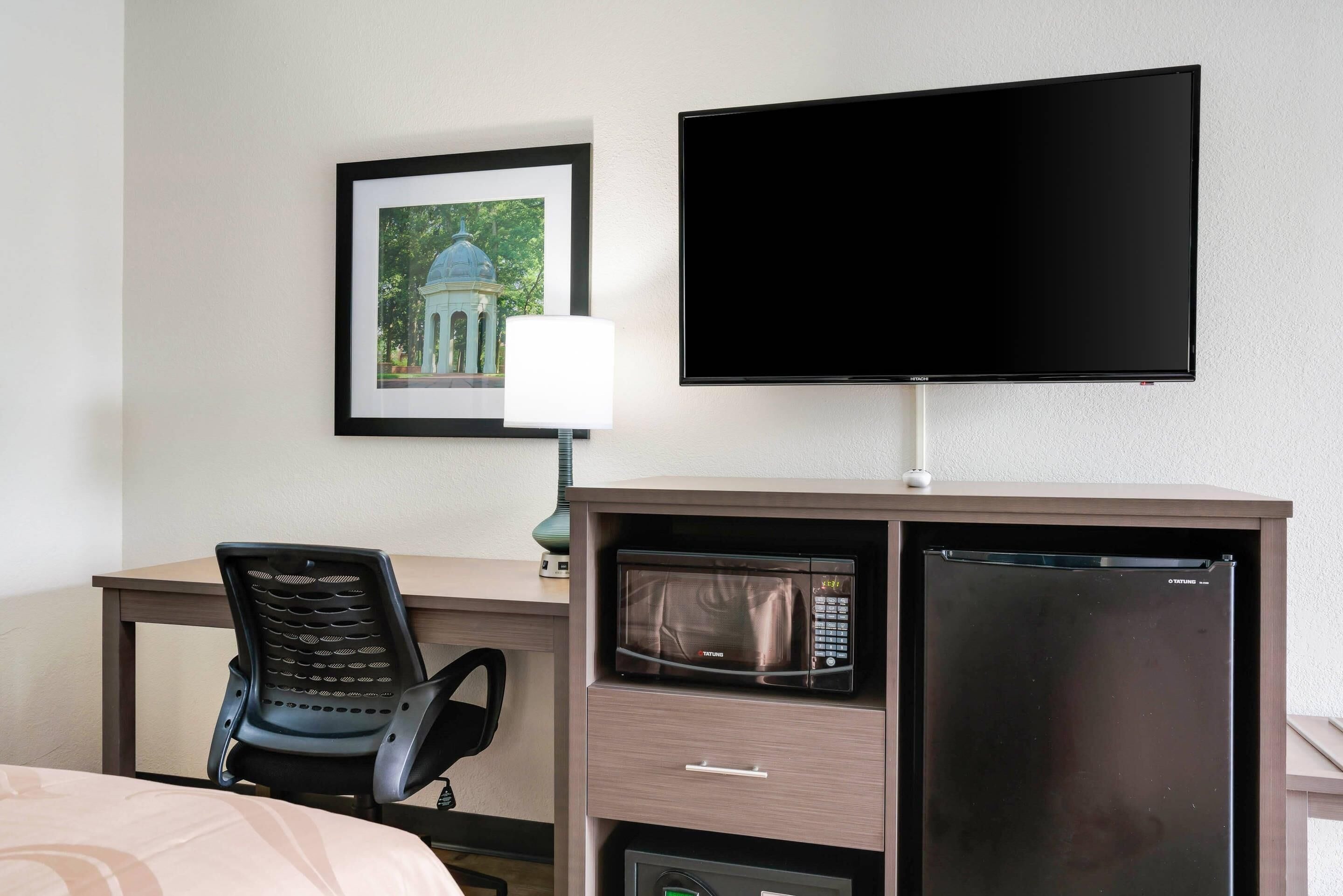 A hotel room with a bed, a desk with a chair, a flat-screen TV, a microwave, and a refrigerator.