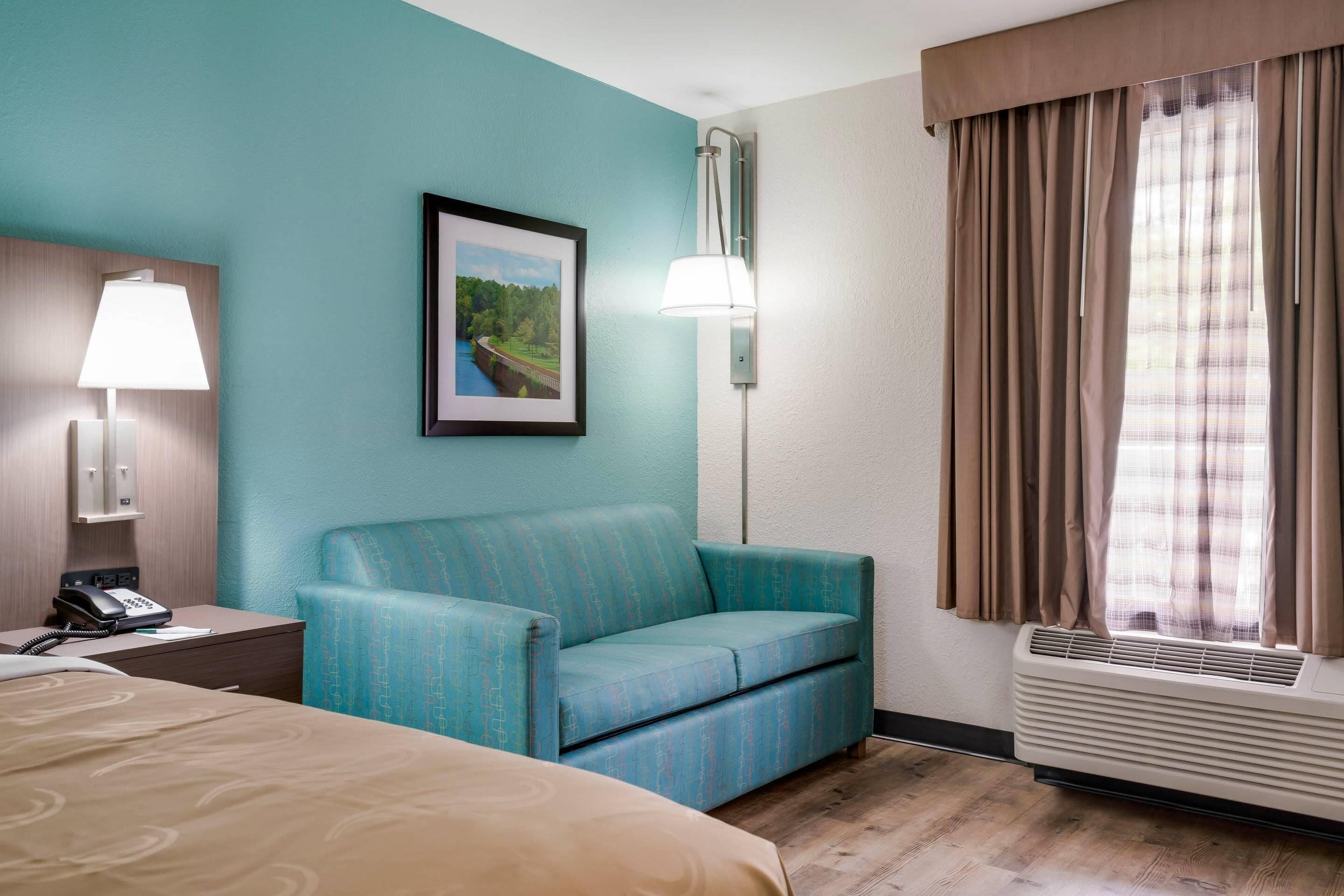 A hotel room with a bed, a desk with a telephone, a blue sofa, a framed picture on the wall, a wall-mounted lamp, and a window with curtains.