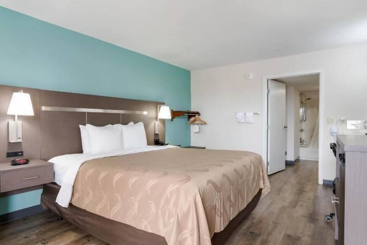A hotel room with a bed, bedside tables, a lamp, a chair, and a bathroom visible through an open door.