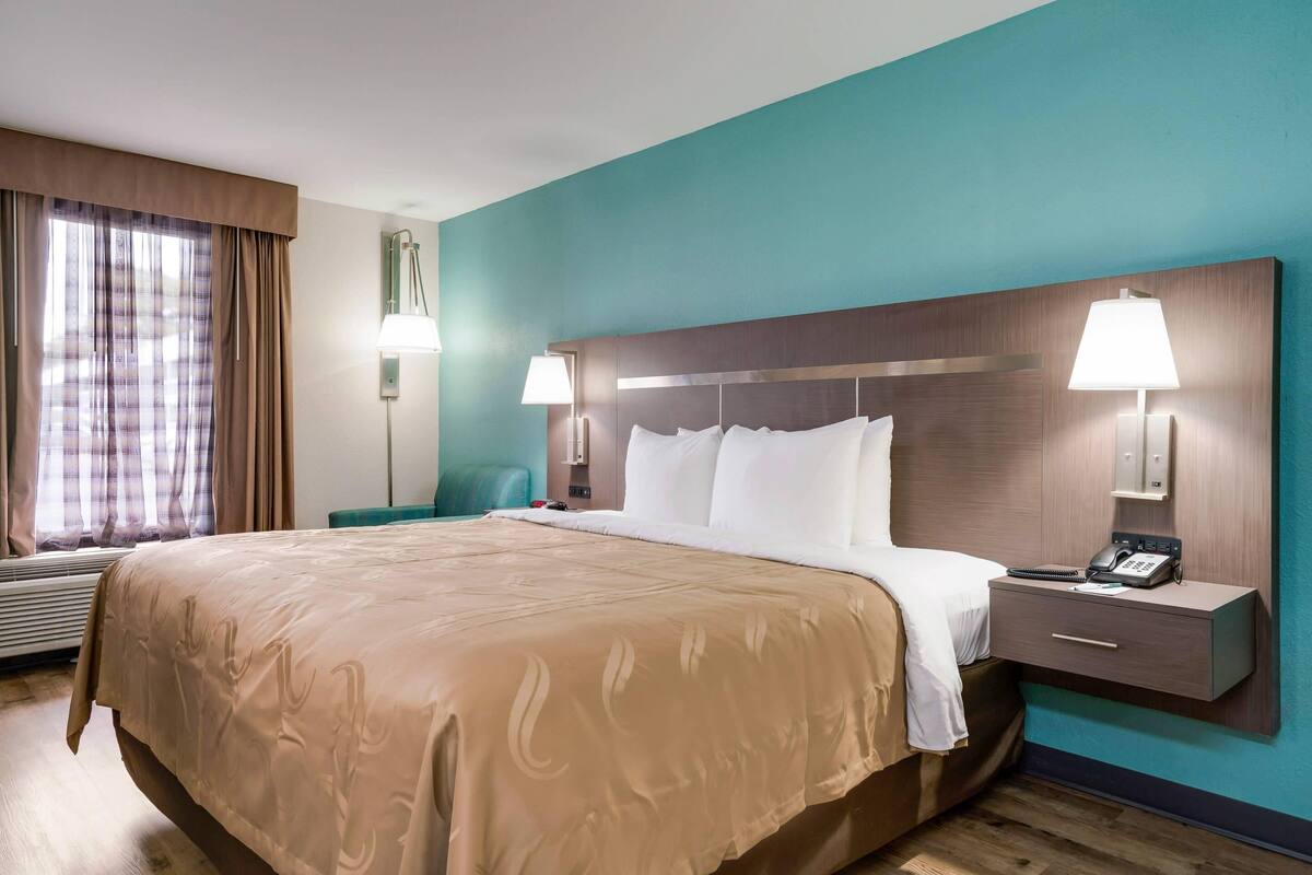 A hotel room with a large bed, two bedside lamps, a nightstand with a phone, and a window with curtains.