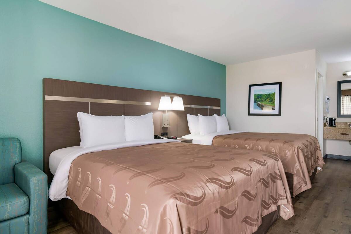A hotel room with two beds, a nightstand, a lamp, a framed picture on the wall, and a chair.