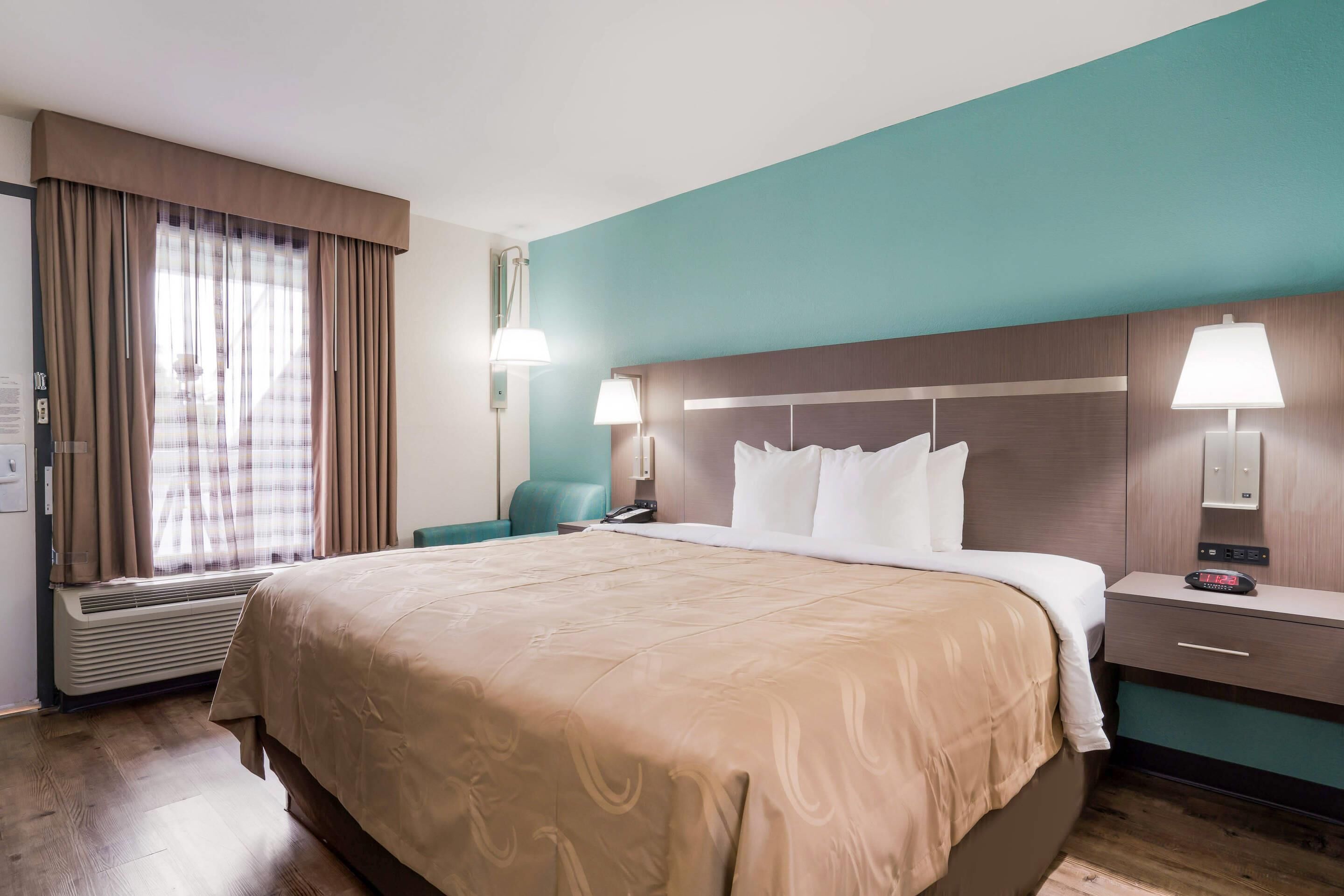Une chambre d’hôtel avec un grand lit, un bureau avec une lampe, une chaise et une fenêtre avec des rideaux.