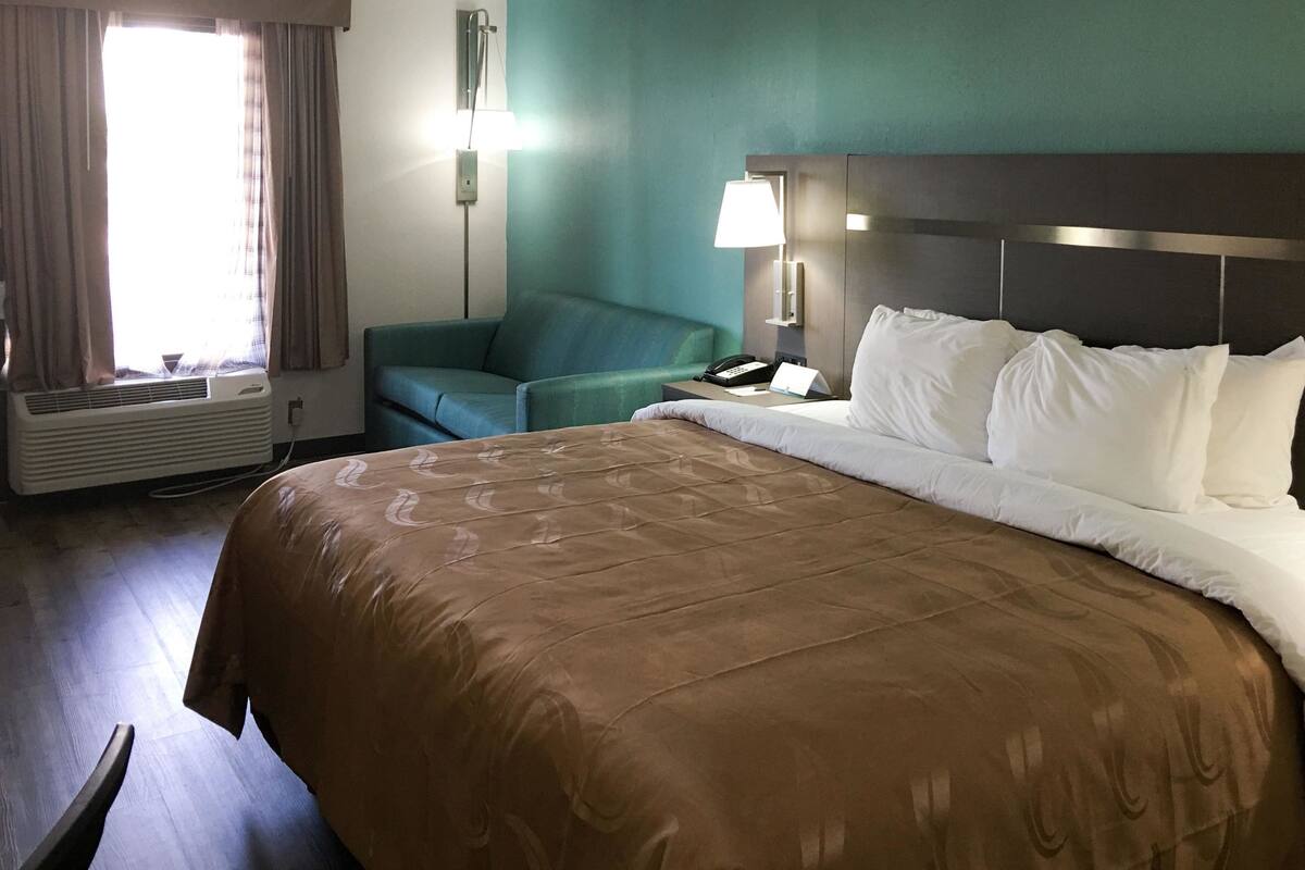 A hotel room with a large bed, a desk with a lamp, a chair, and a window with curtains.