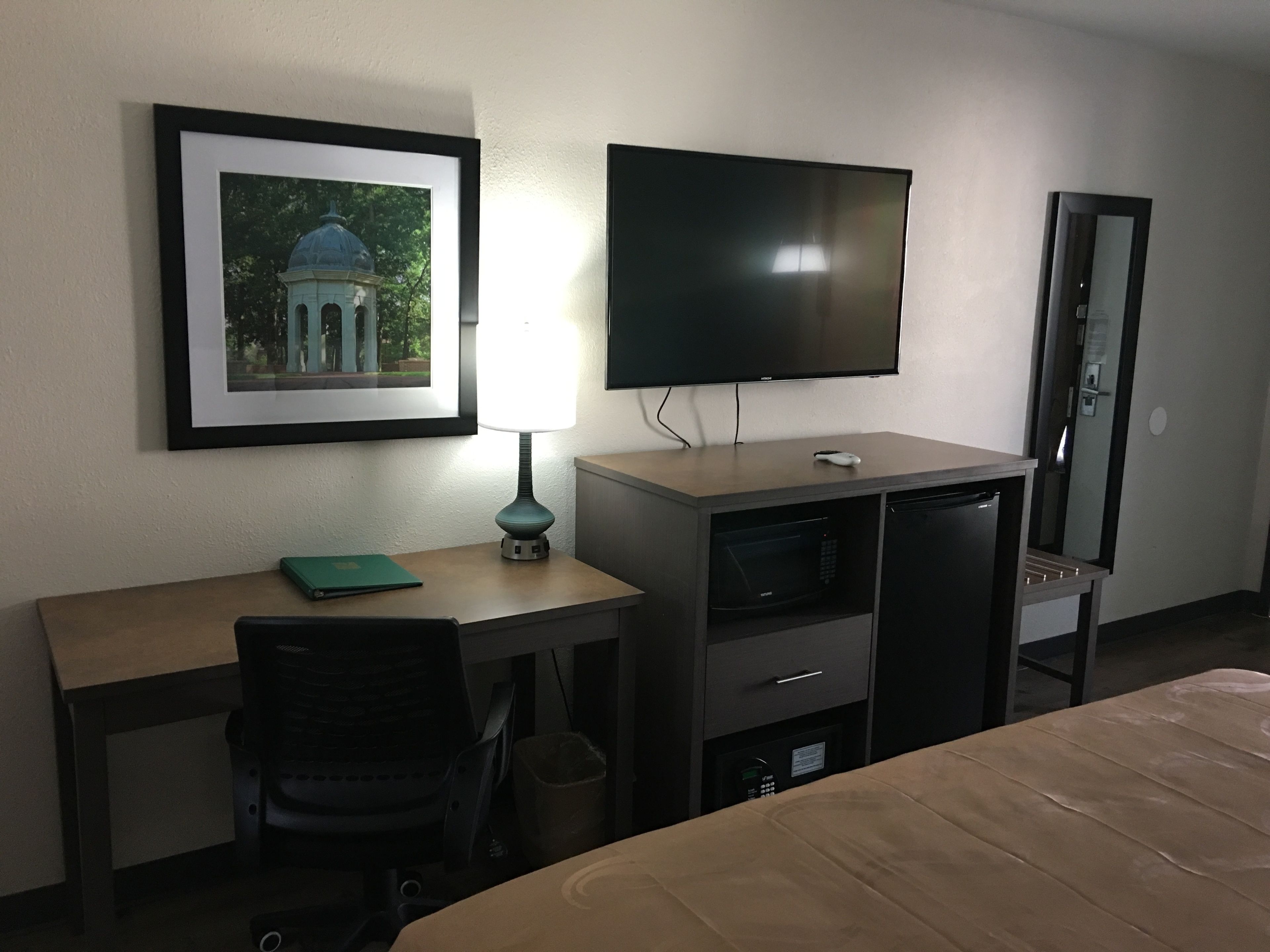 A hotel room with a bed, a desk with a chair, a TV mounted on the wall, a framed picture, and a lamp.