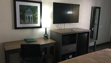 A hotel room with a bed, a desk with a chair, a TV mounted on the wall, a framed picture, and a lamp.