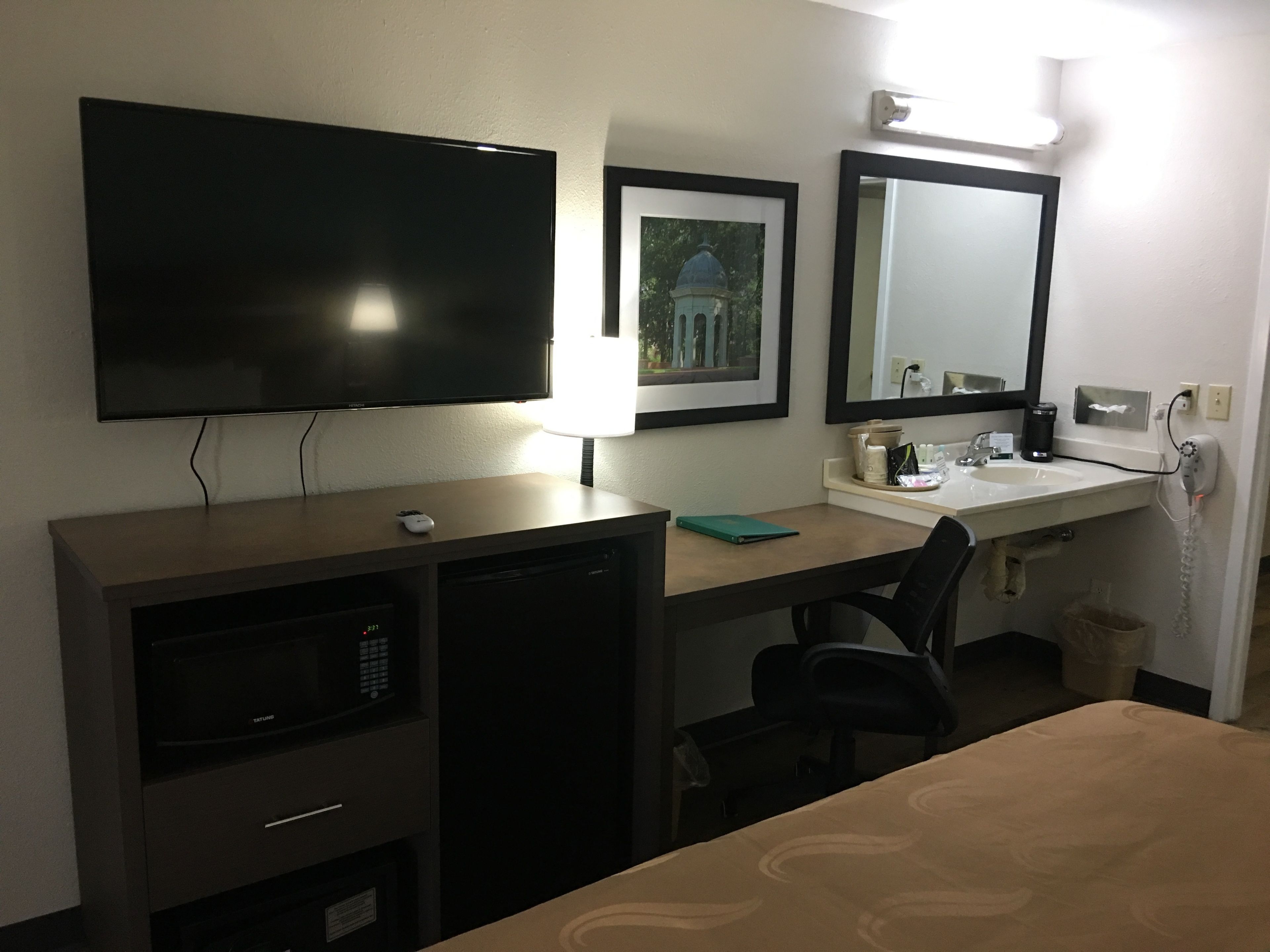 Hotelkamer met flatscreen televisie, bureau met lamp, spiegel, wastafel, haardroger en een klein prullenbakje.